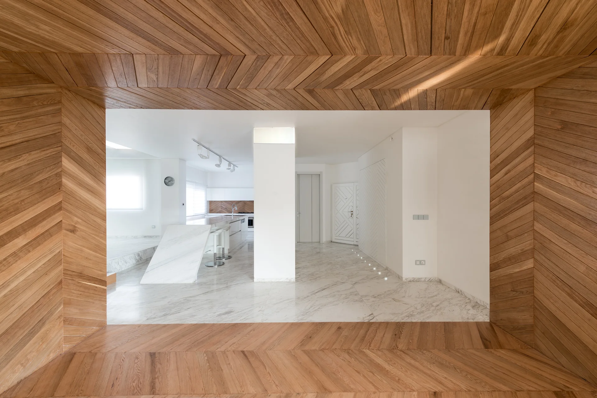A geometric wood-paneled archway frames a view of a minimalist white kitchen featuring a sculptural marble island and marble flooring.