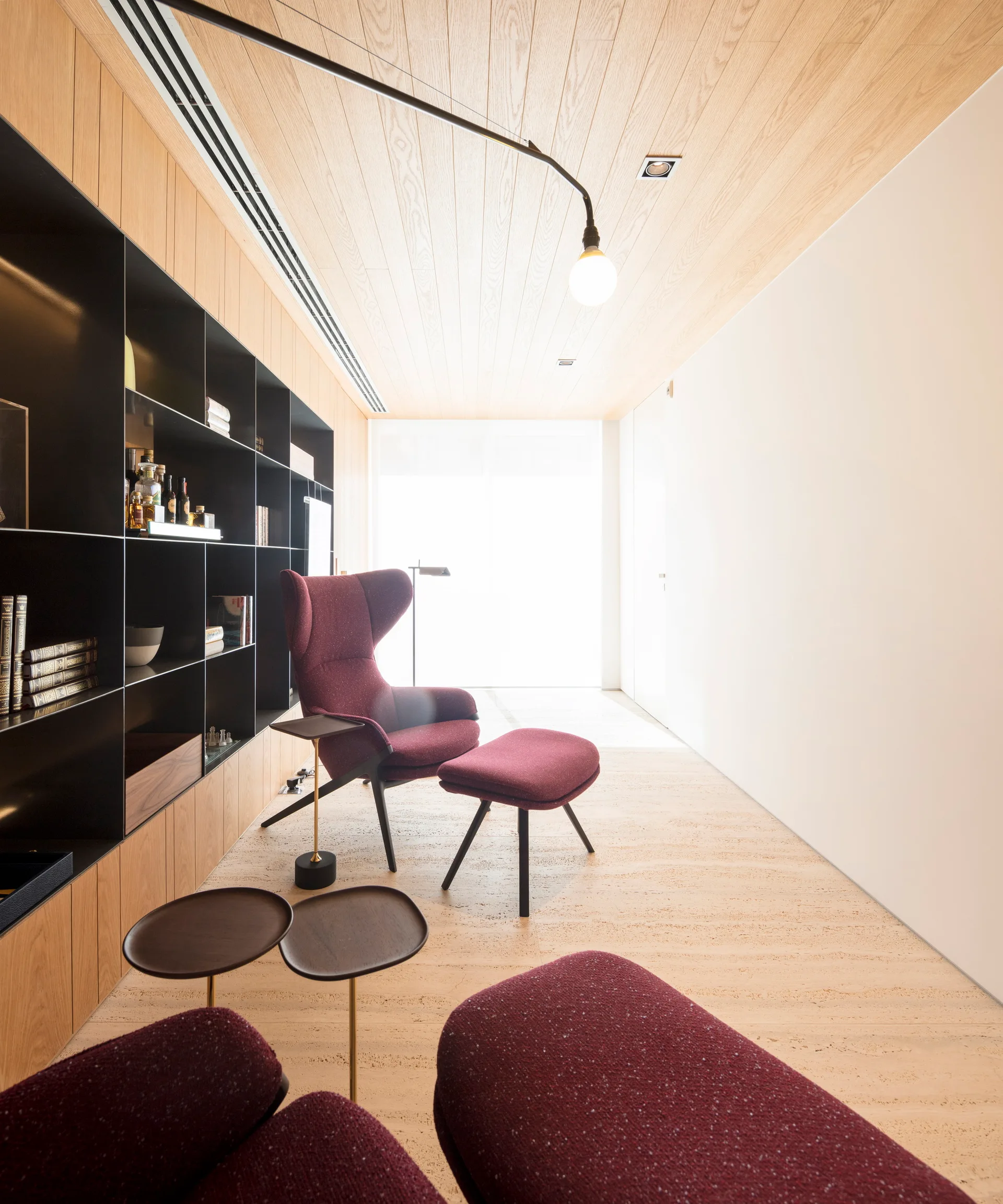 A modern lounge room features a burgundy winged armchair with an ottoman next to a large black built-in shelving unit under a wood plank ceiling.