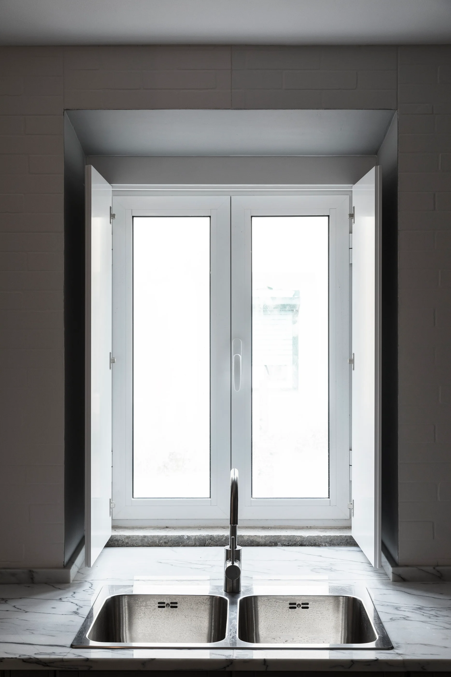 A stainless steel double-basin kitchen sink is set in a veined marble countertop directly beneath a window with open white shutters.