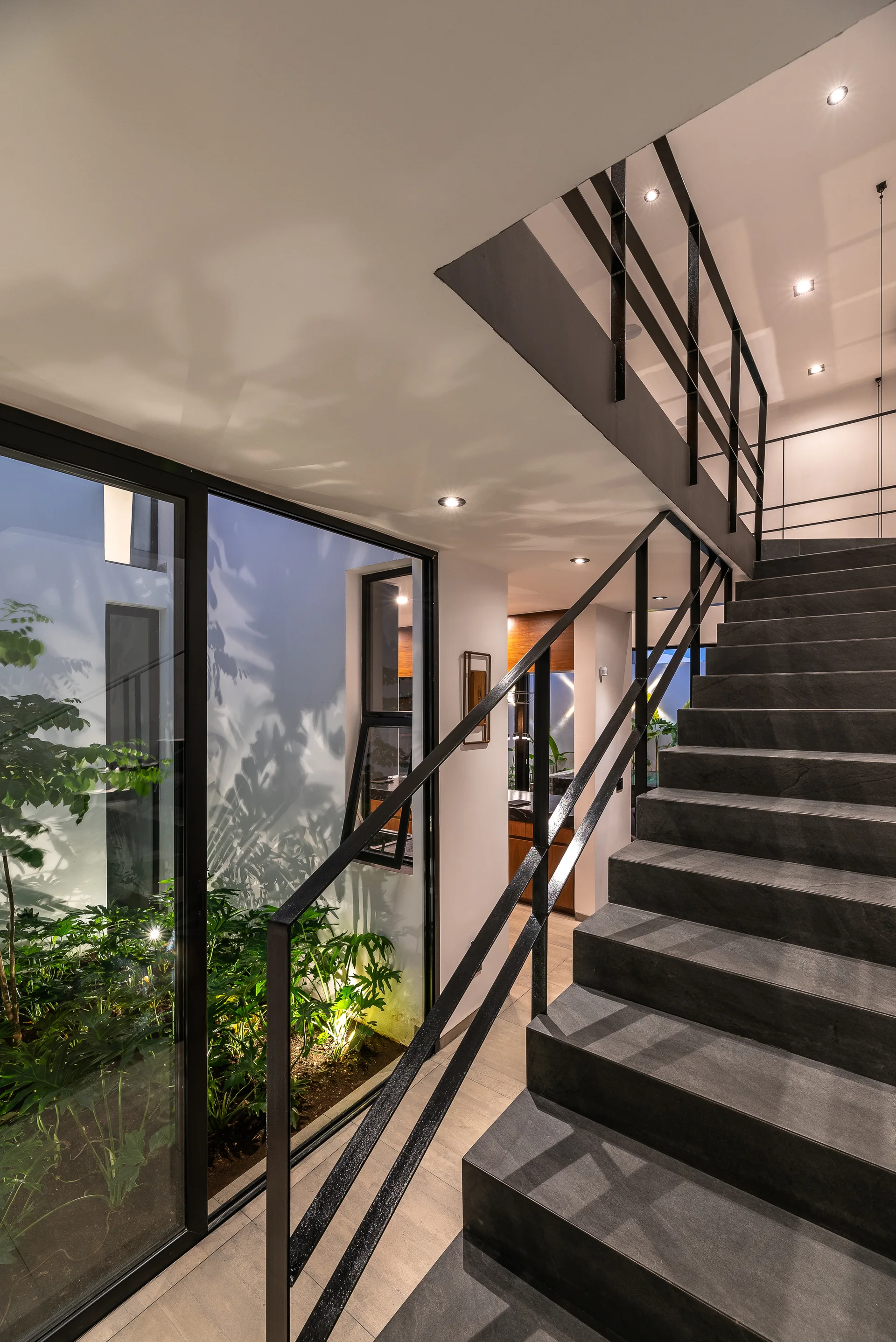 A modern staircase features dark stone steps and a black railing next to large glass windows looking out onto a lit garden courtyard.