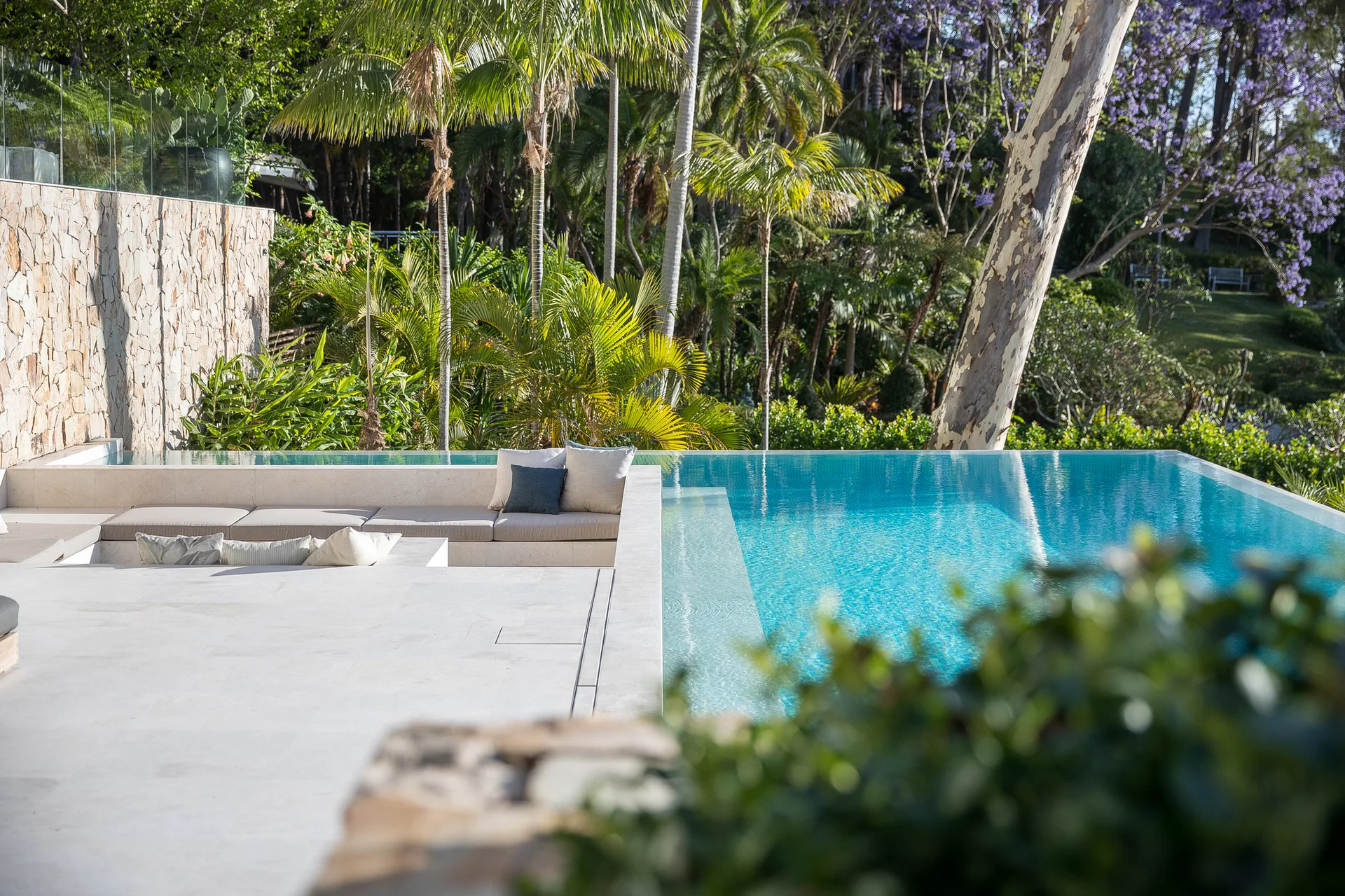 An outdoor infinity pool with clear blue water features a sunken stone seating area with light cushions, set against a backdrop of lush green palm trees.