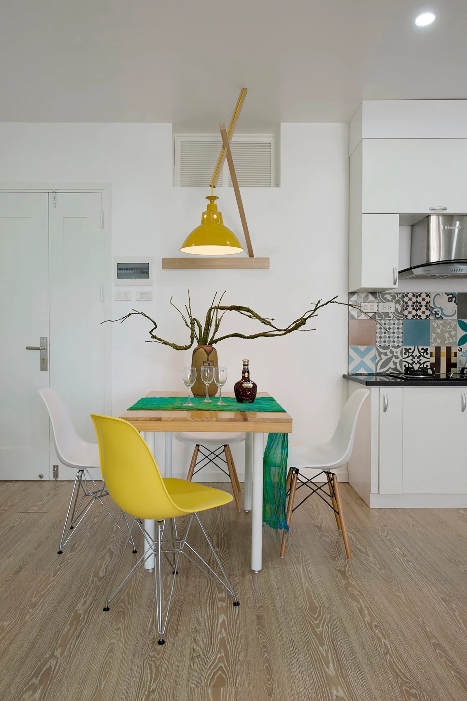 A small wooden dining table is paired with white and yellow shell chairs under a yellow pendant light, next to a kitchen with patterned tile backsplash.