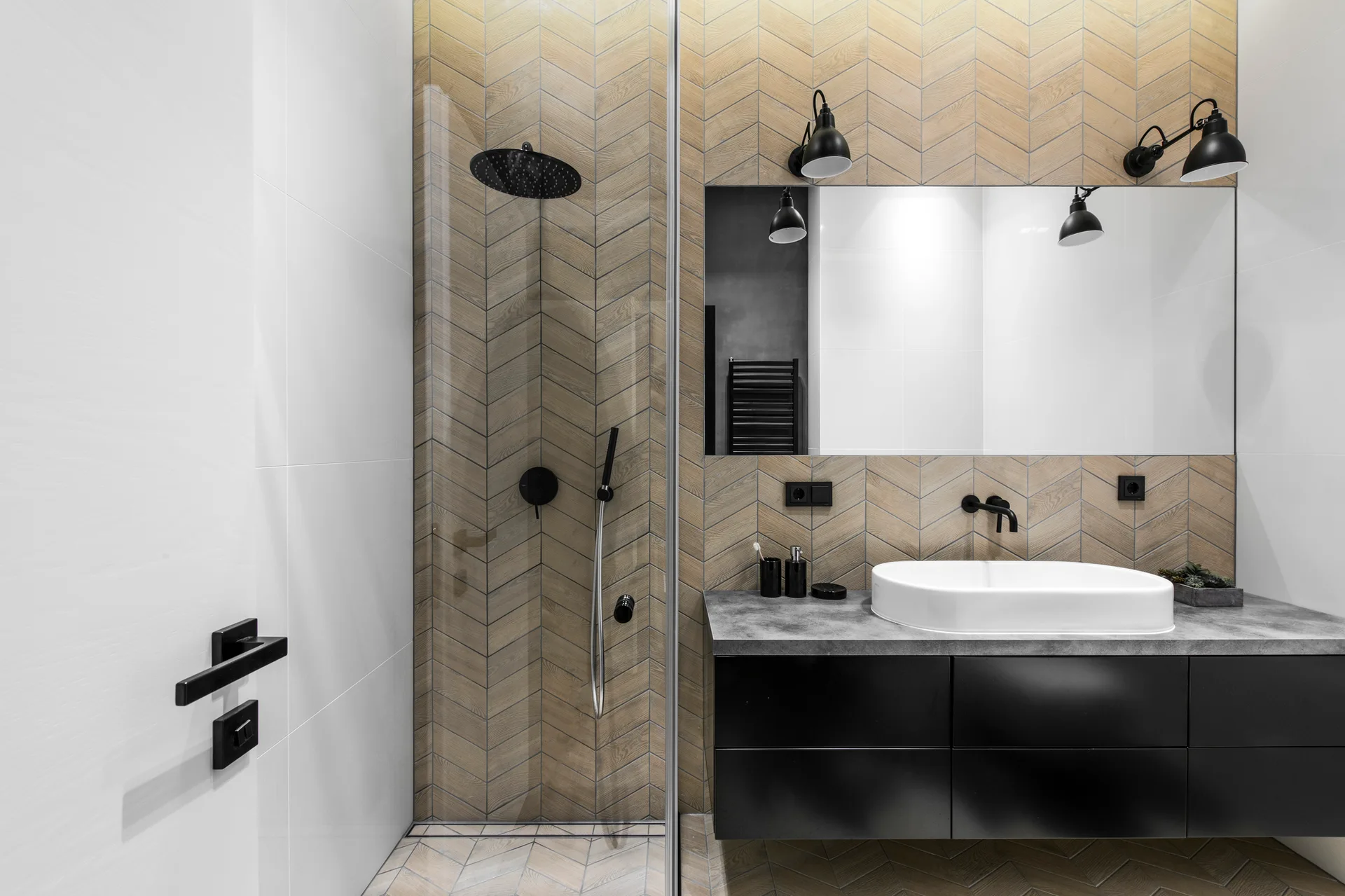 A modern bathroom featuring a black floating vanity with a white vessel sink, a large mirror, and wood-look chevron tile walls and floor.
