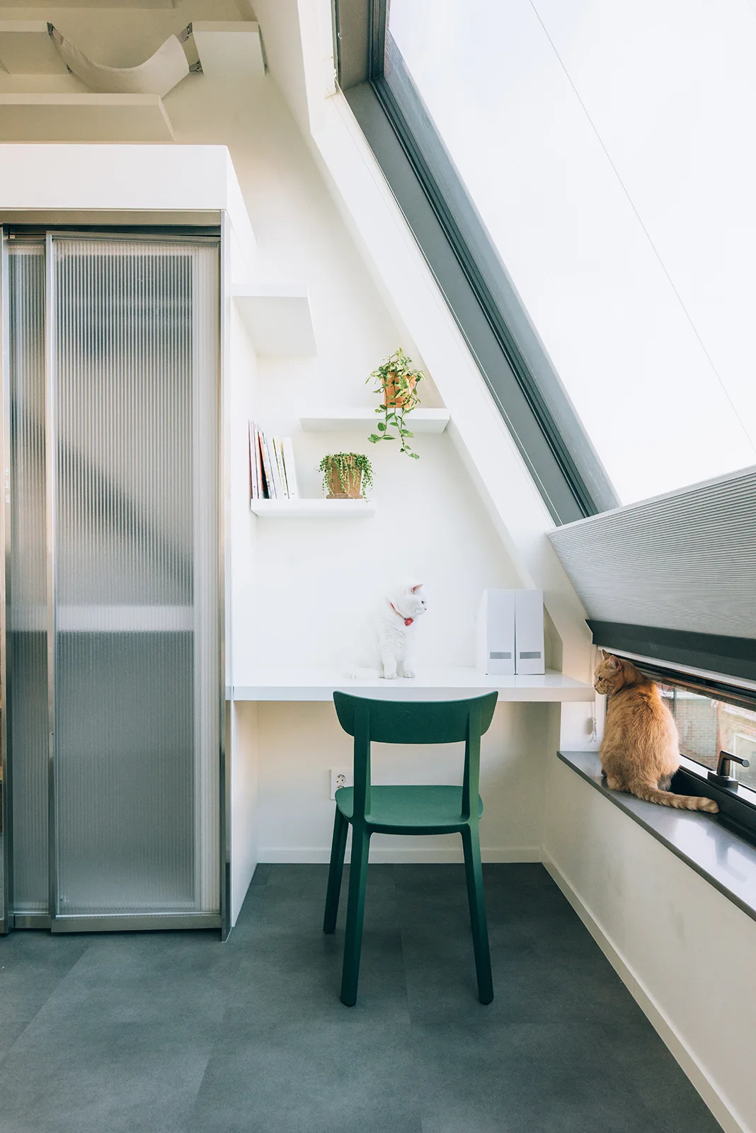A minimalist home office with a built-in white desk, a green chair, floating shelves, and a large sloped window where two cats are sitting.