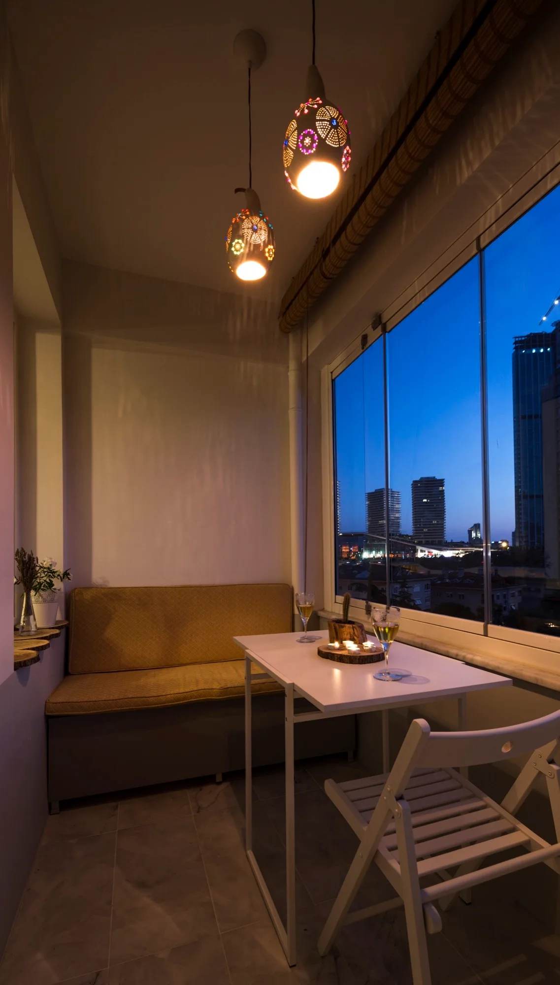 An enclosed balcony dining area with a mustard bench, small white table, and two patterned glowing pendant lights overlooking a city skyline at dusk.
