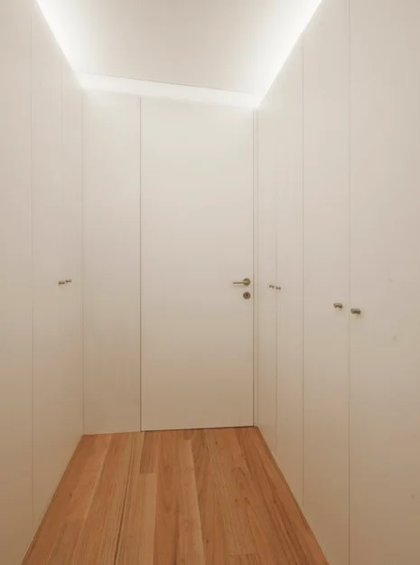 A narrow closet space featuring floor-to-ceiling white cabinetry, wood plank flooring, and soft indirect cove lighting along the ceiling.
