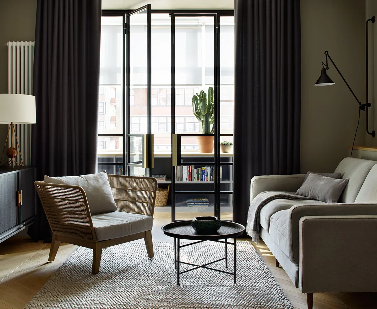 A light gray sofa and a woven-backed armchair frame a black metal coffee table on a textured rug in front of tall glass-paneled doors with dark drapes.
