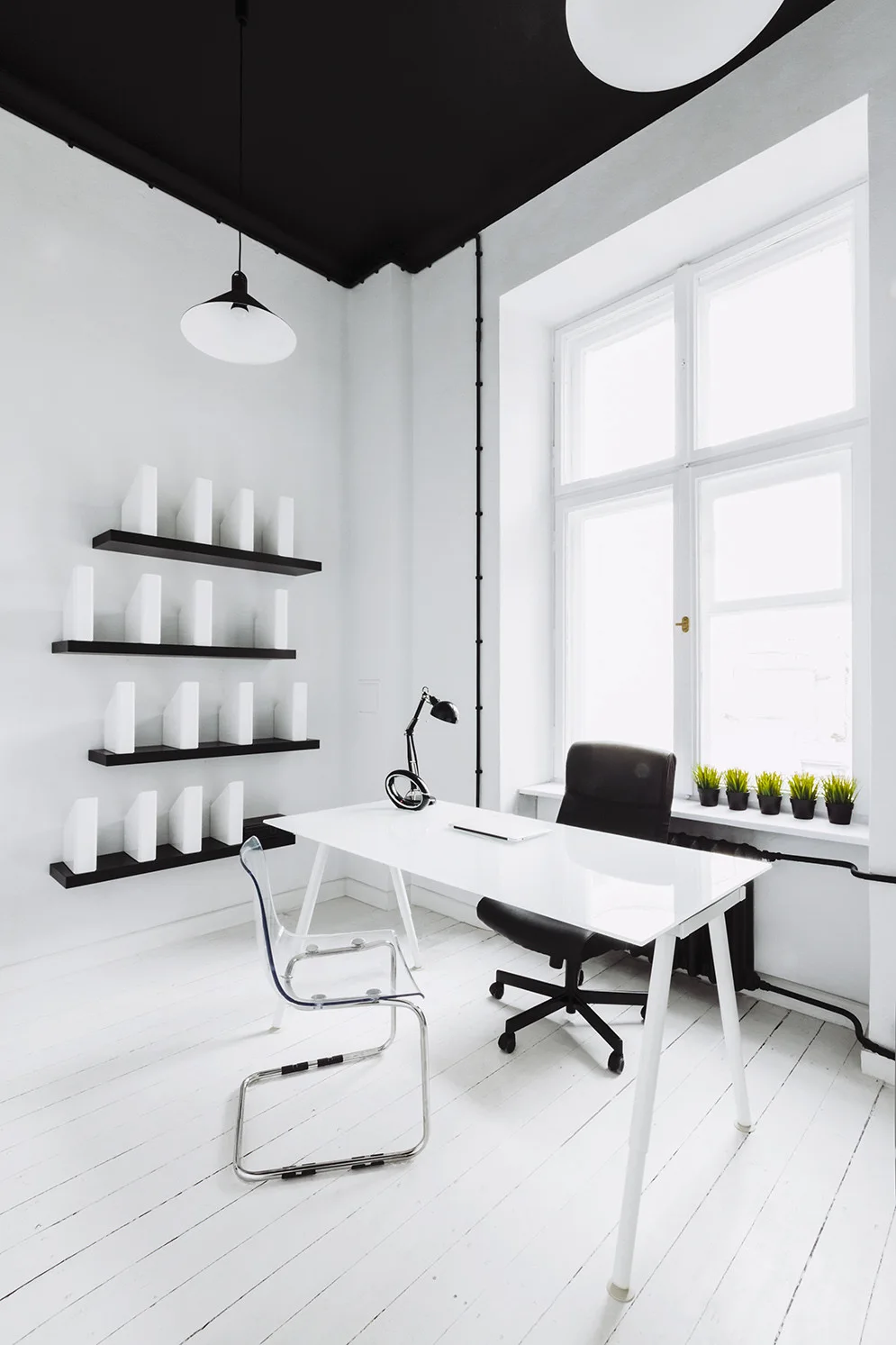 A minimalist home office features a white desk, a black office chair, open floating black shelves, and a striking black painted ceiling.