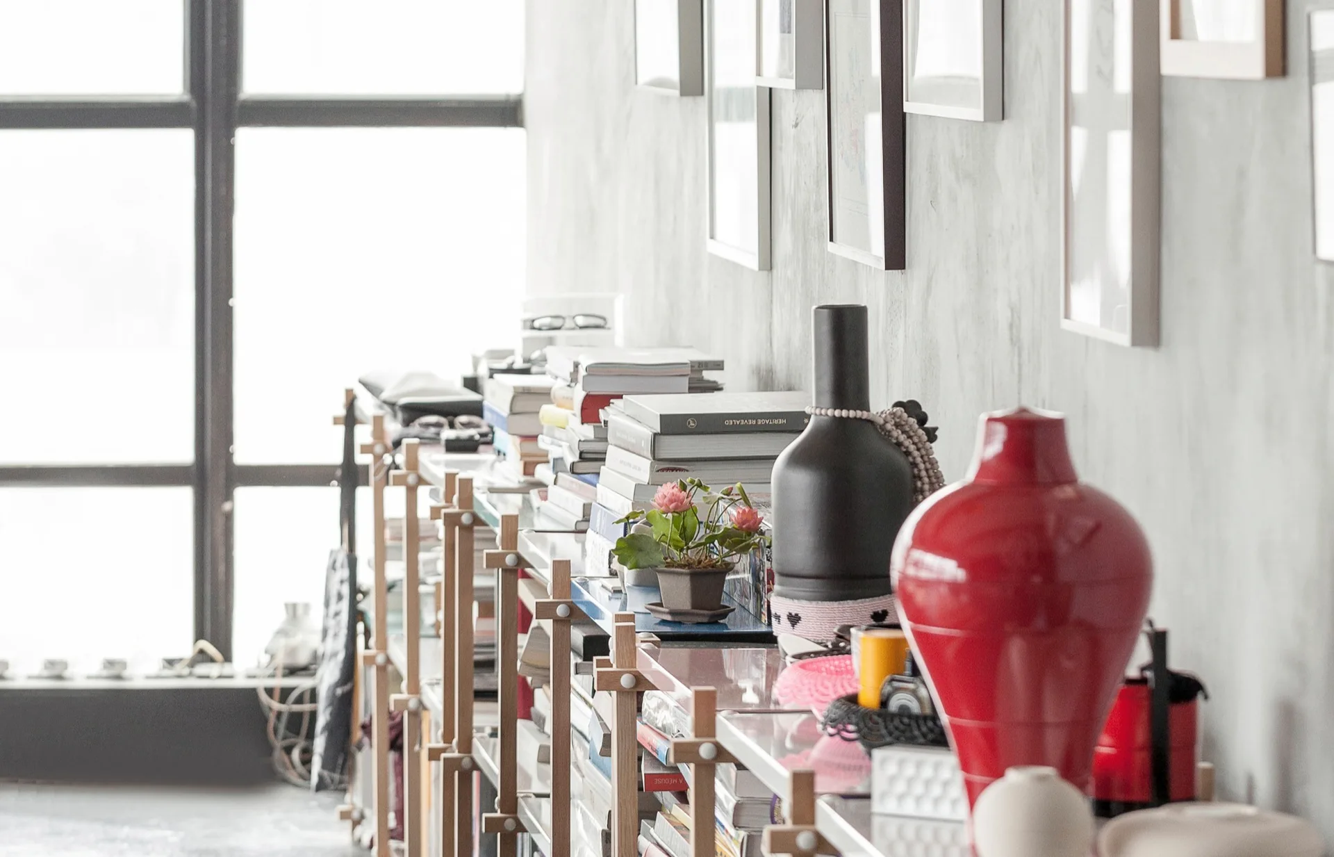 A low wooden shelving unit filled with stacks of books, a vibrant red vase, and a black vase against a grey wall with framed art.