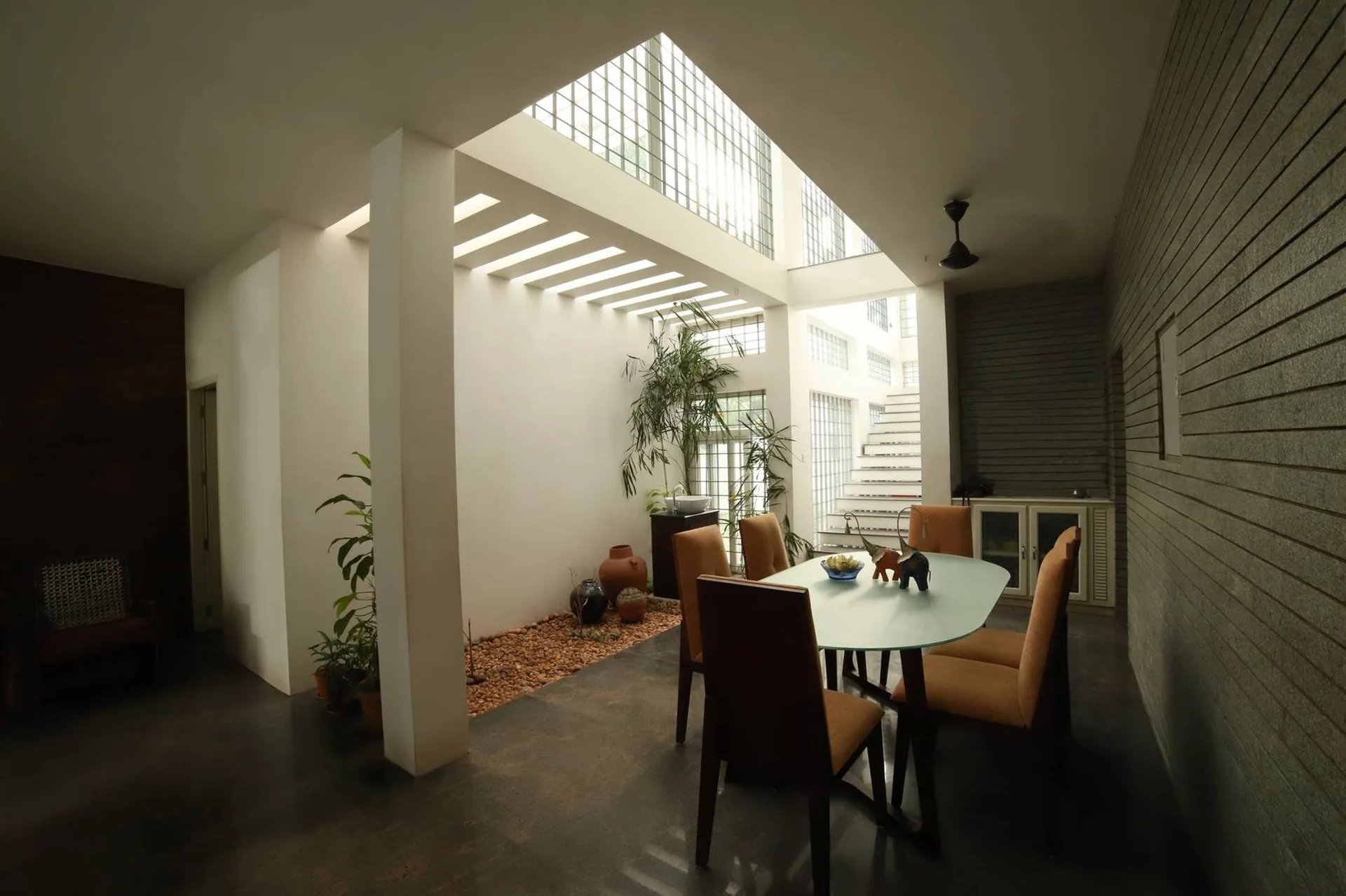 A modern dining room features a glass-top table with tan chairs beneath a tall skylight atrium and a small indoor pebble bed.