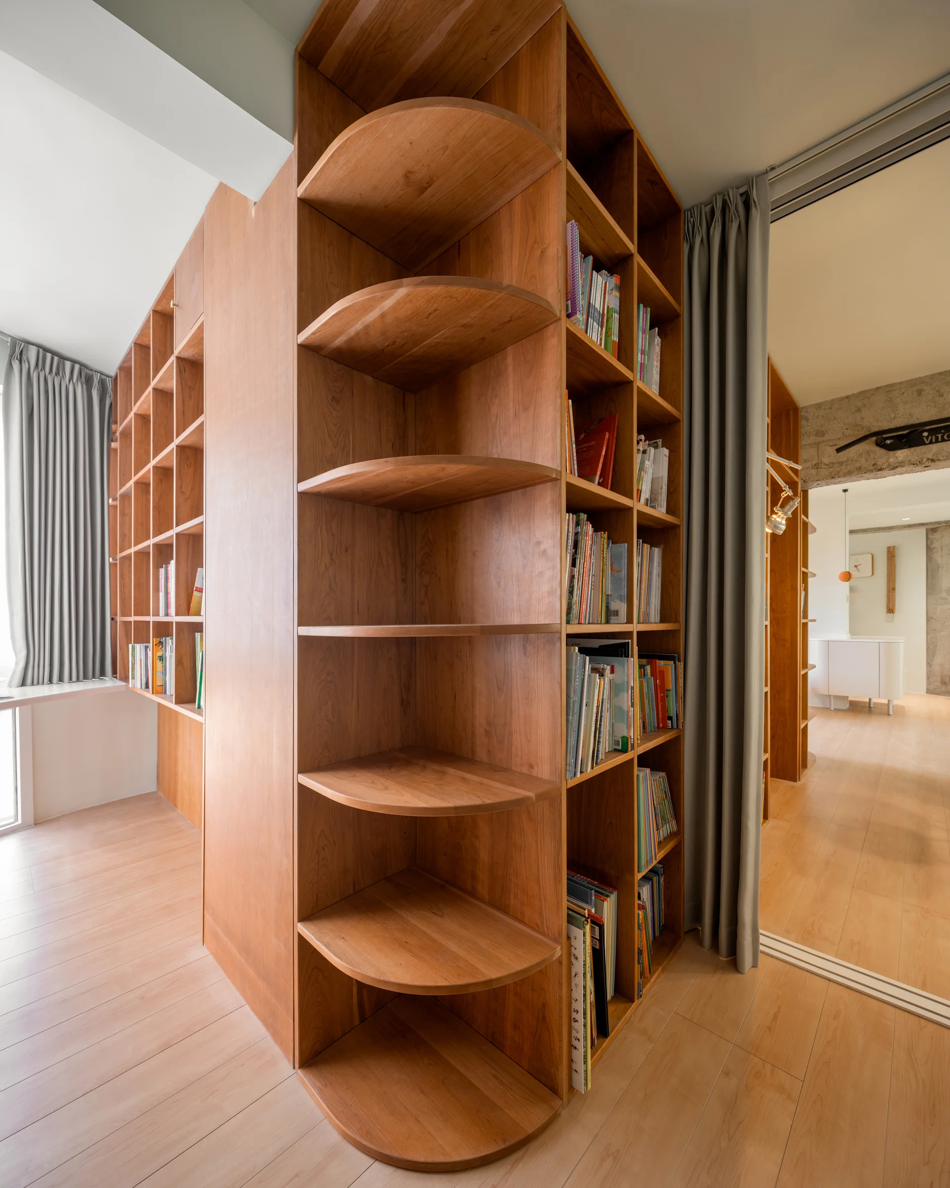 A tall wood bookshelf with curved outer shelves acts as a room divider next to light wood flooring and grey drapes.