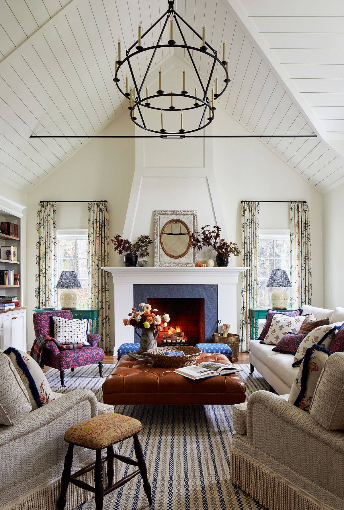 A traditional living room features a high vaulted plank ceiling, a central fireplace with a mantel, and a tufted leather ottoman on a striped rug.