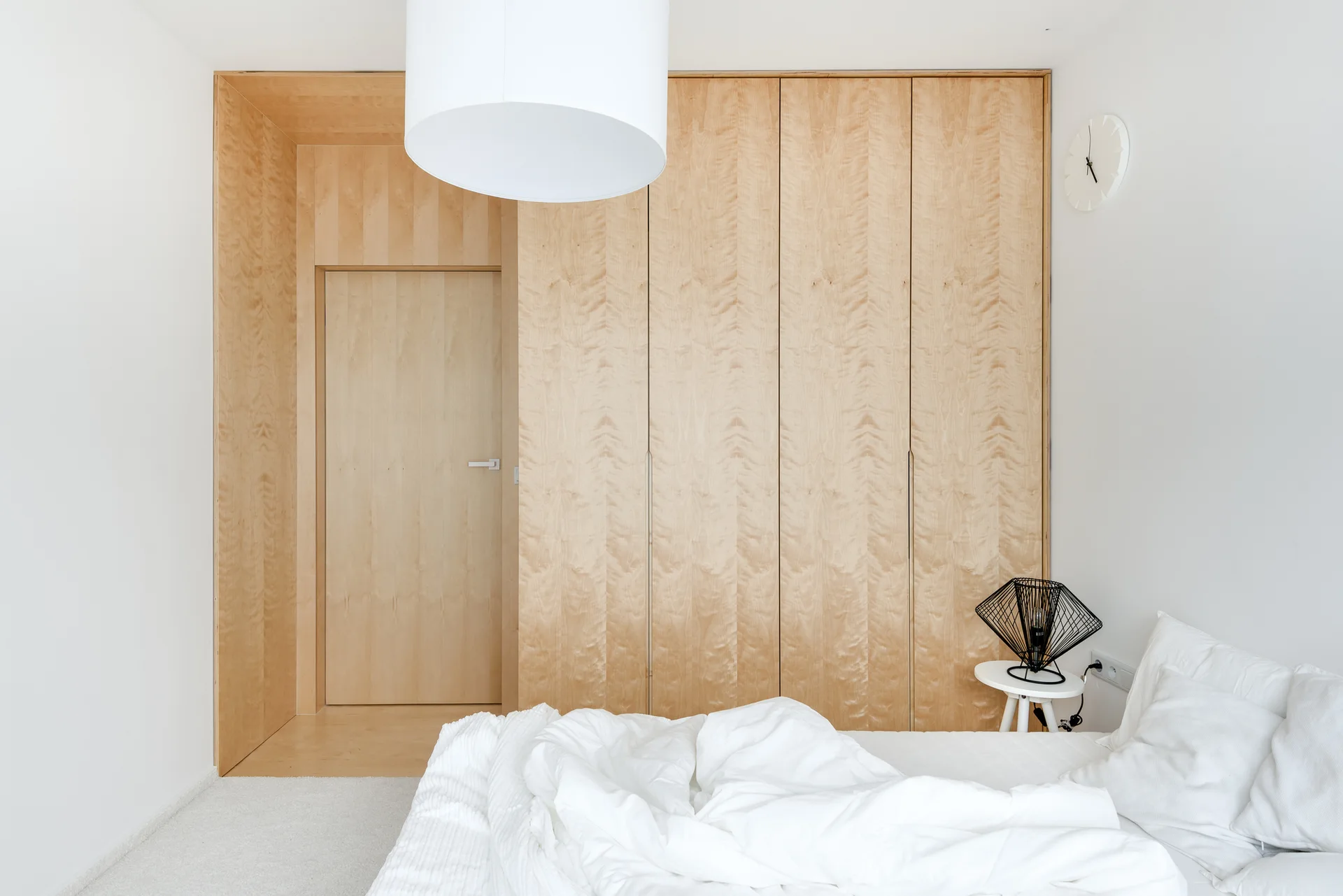 A minimalist bedroom featuring white bedding, a large white drum pendant, and a floor-to-ceiling light wood cabinetry wall with an integrated flush door.