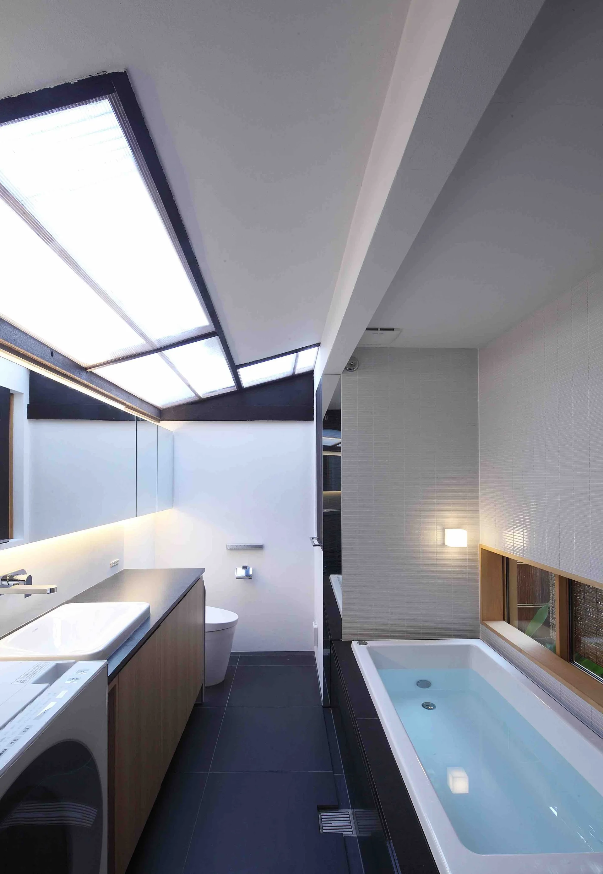 A minimalist bathroom with a sloped glass skylight, a built-in tub with a low window, and a wood-toned vanity on dark tile flooring.