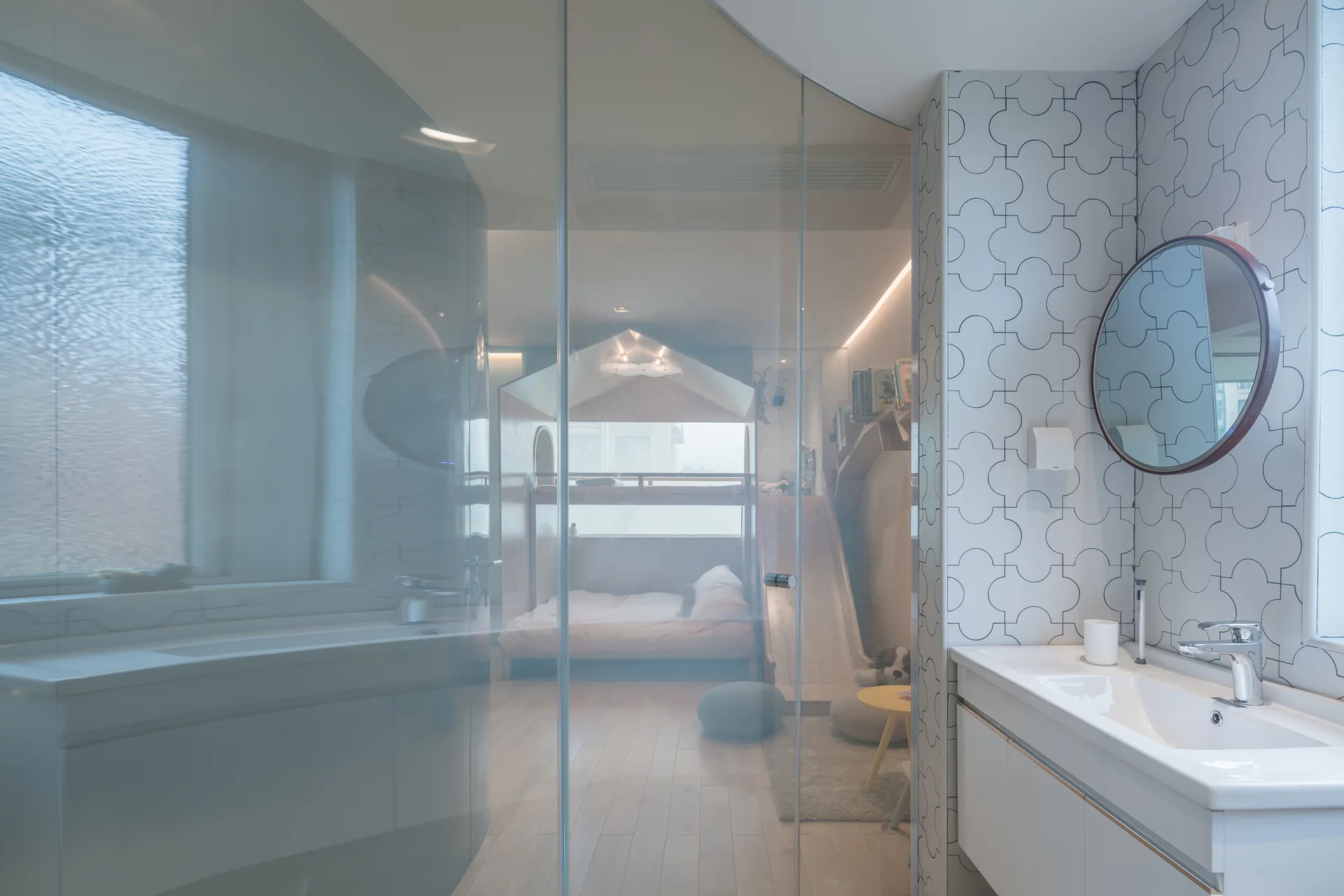 A modern bathroom with a white vanity and patterned tiles features a curved glass partition wall overlooking a children's bedroom.