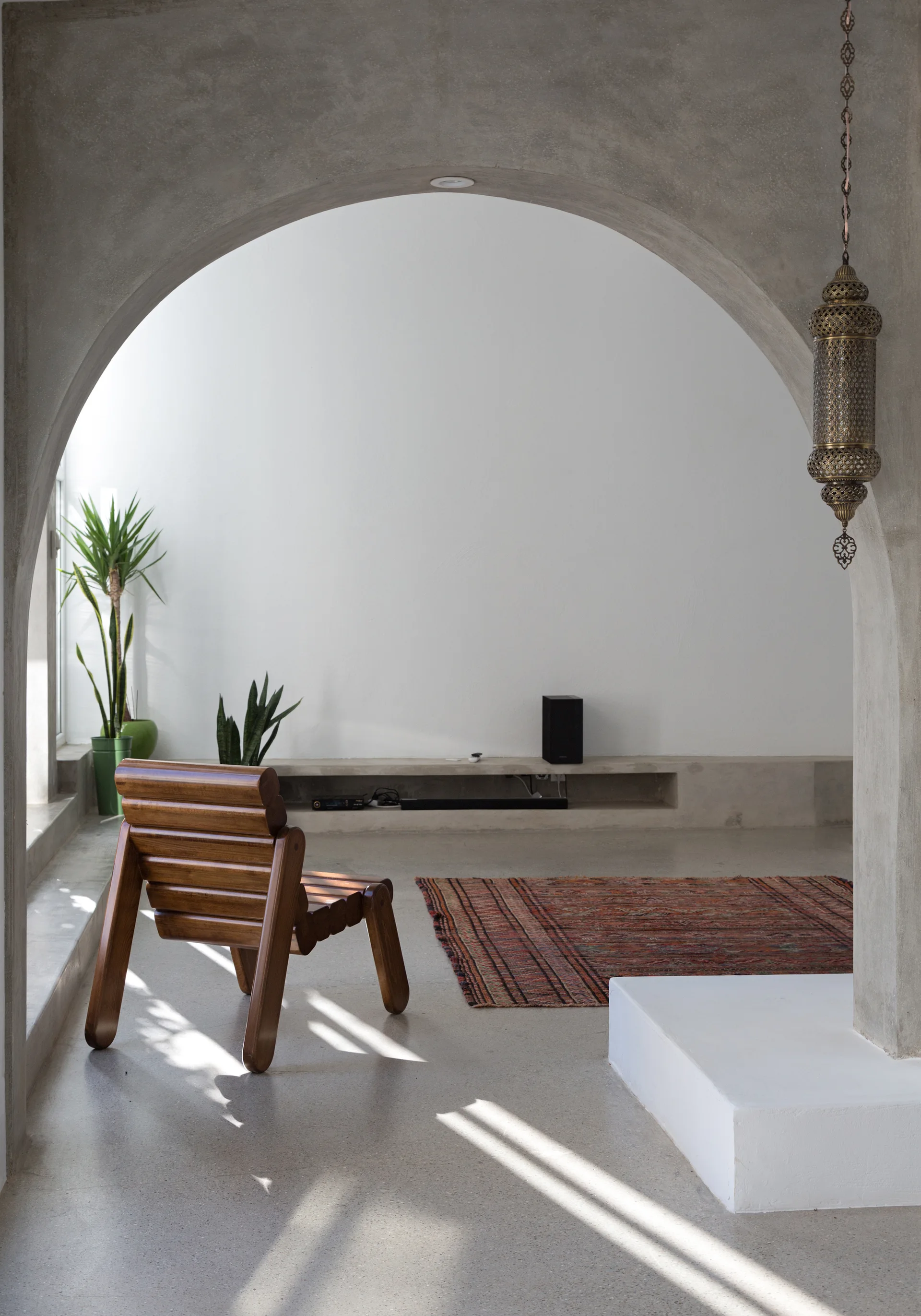 A concrete archway frames a minimalist living room with white plaster walls, a wooden armchair, a patterned rug, and a hanging metal lantern.