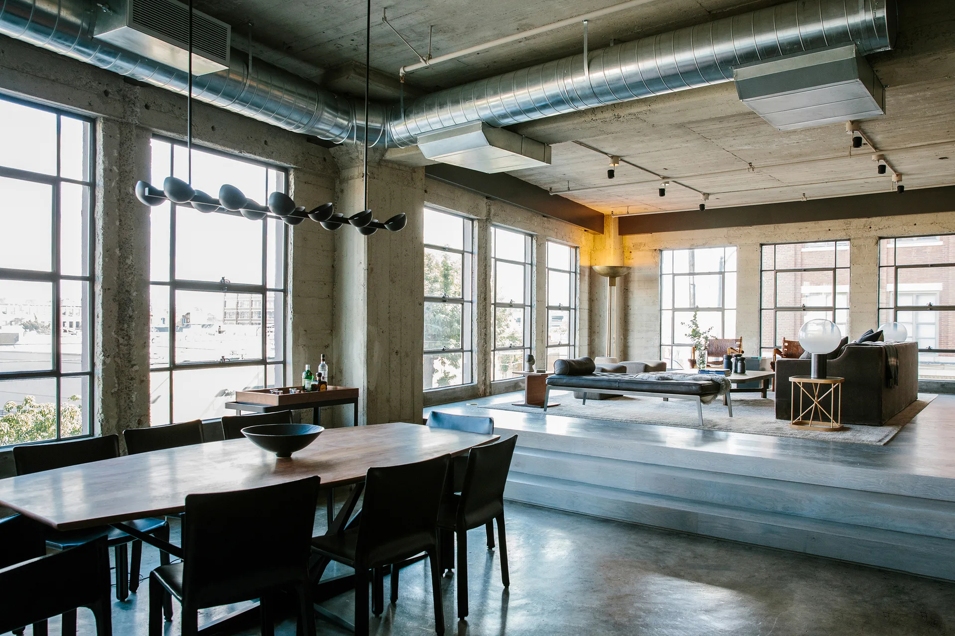 An industrial loft space with a wooden dining table in the foreground and an elevated living area on a wooden platform beneath a concrete ceiling.