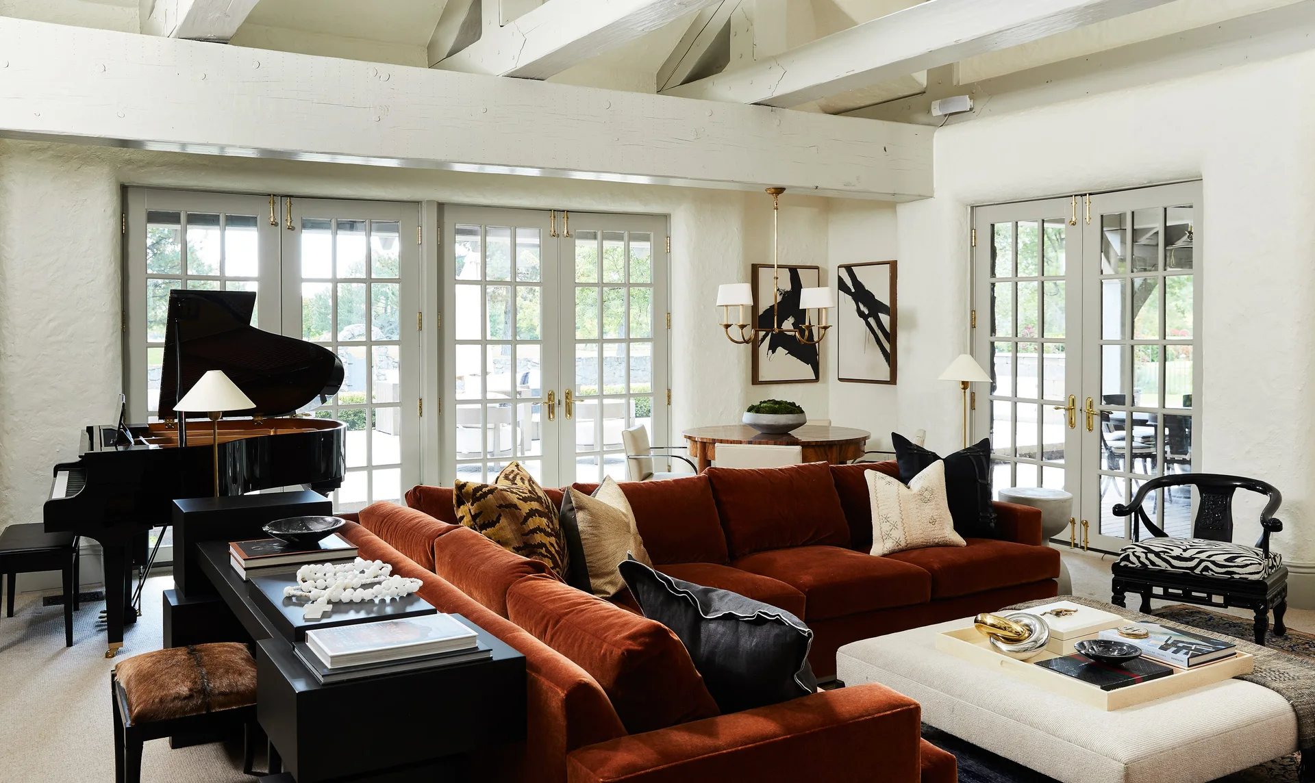 A living room with textured white walls features a rust velvet sectional sofa, a black grand piano, and exposed white ceiling beams.