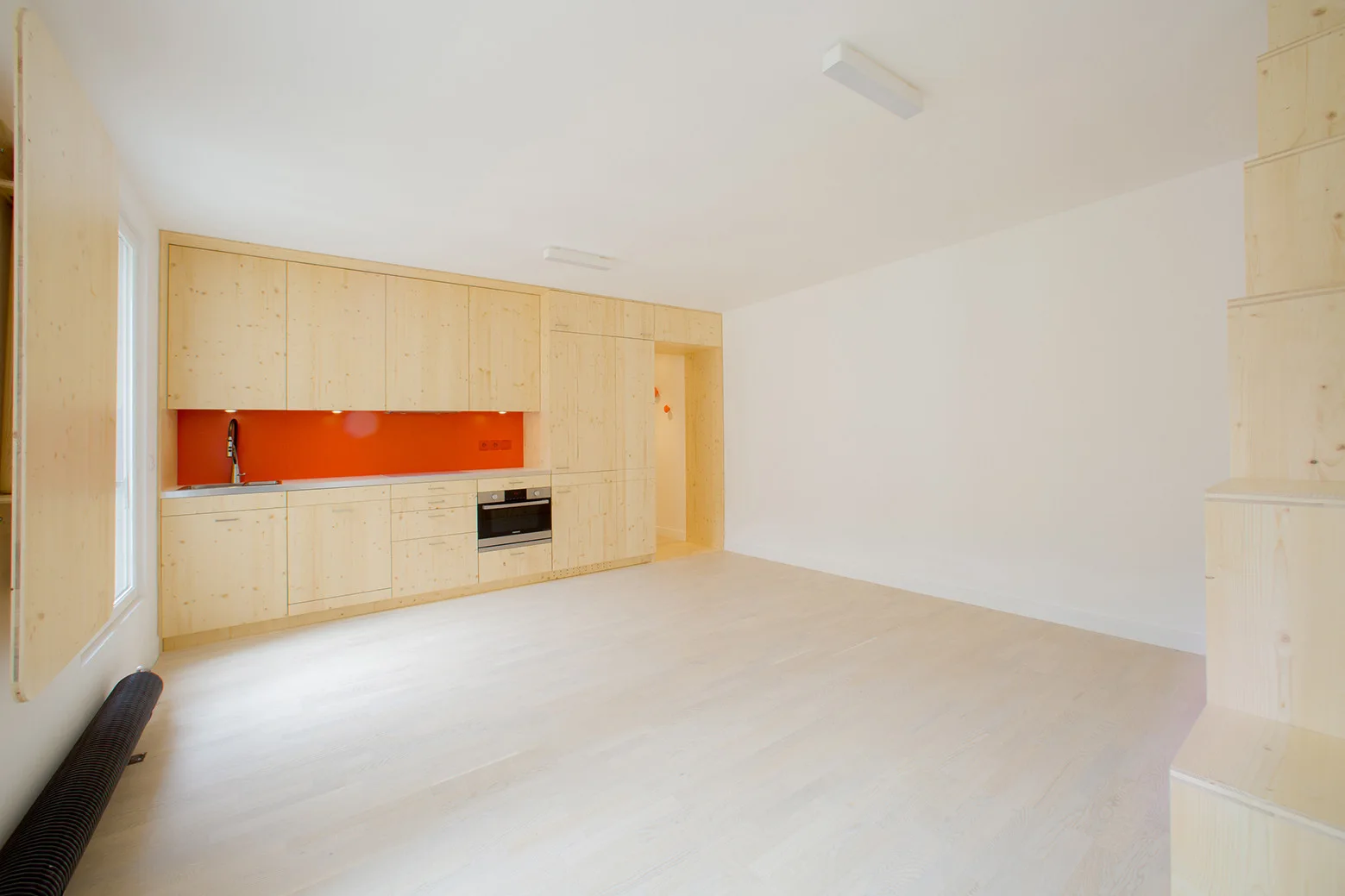 A minimalist kitchen features seamless light wood cabinetry, a bright orange backsplash, pale wood flooring, and a wooden block staircase to the right.