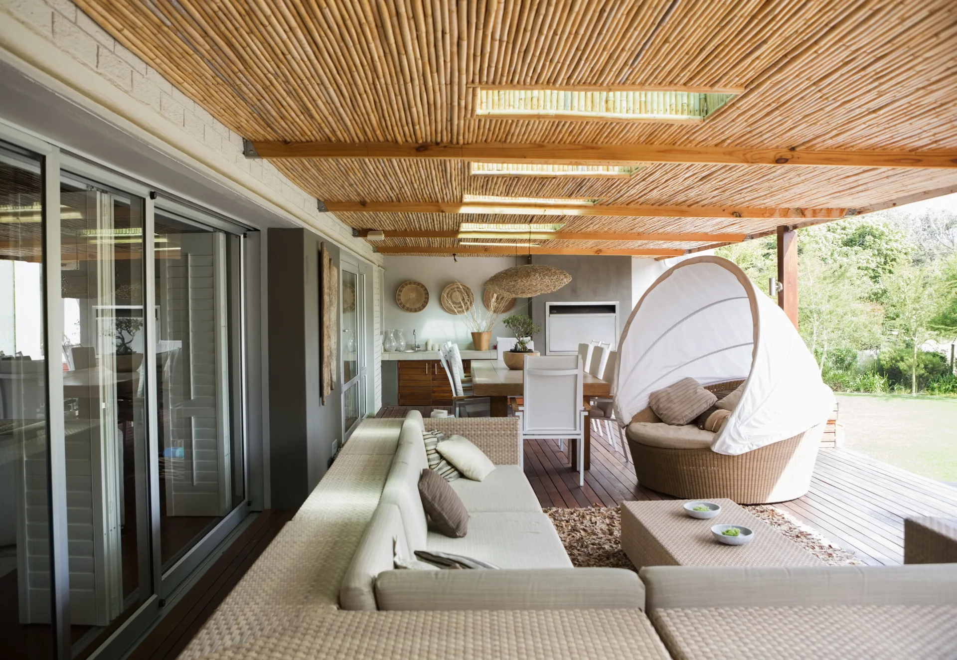 A covered outdoor patio features a slatted bamboo ceiling with skylights, a large wicker sectional sofa, and a matching canopy daybed.