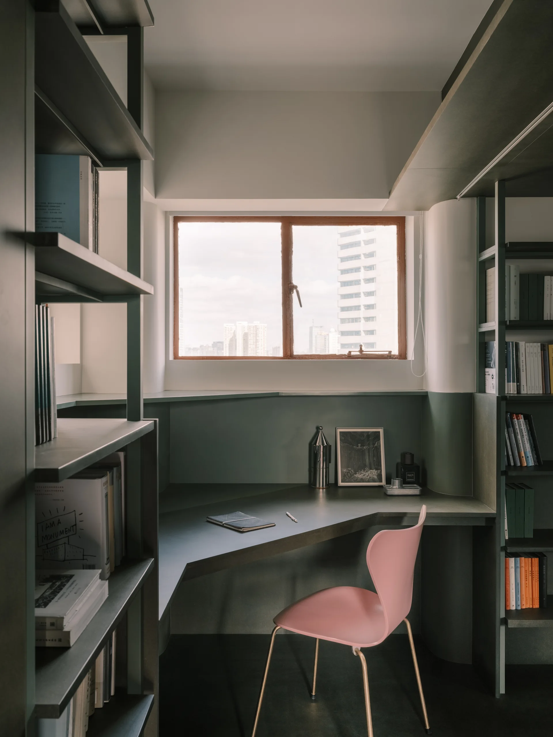 A compact home office nook features a built-in green desk, flanking bookshelves, and a minimalist pink chair placed in front of a window.