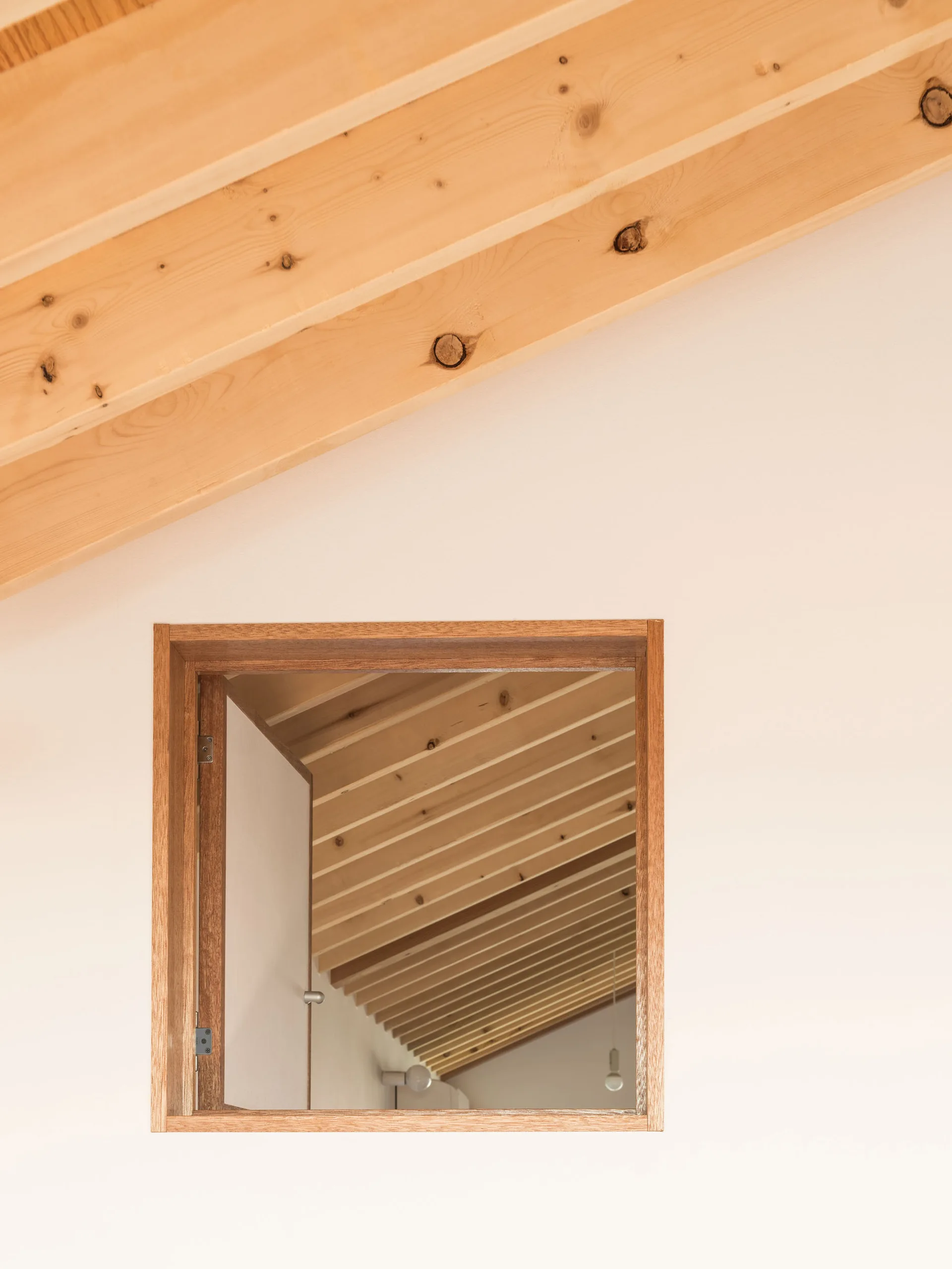 A square wood-framed interior window looks through to a space with a slanted ceiling and exposed wooden beams against white walls.