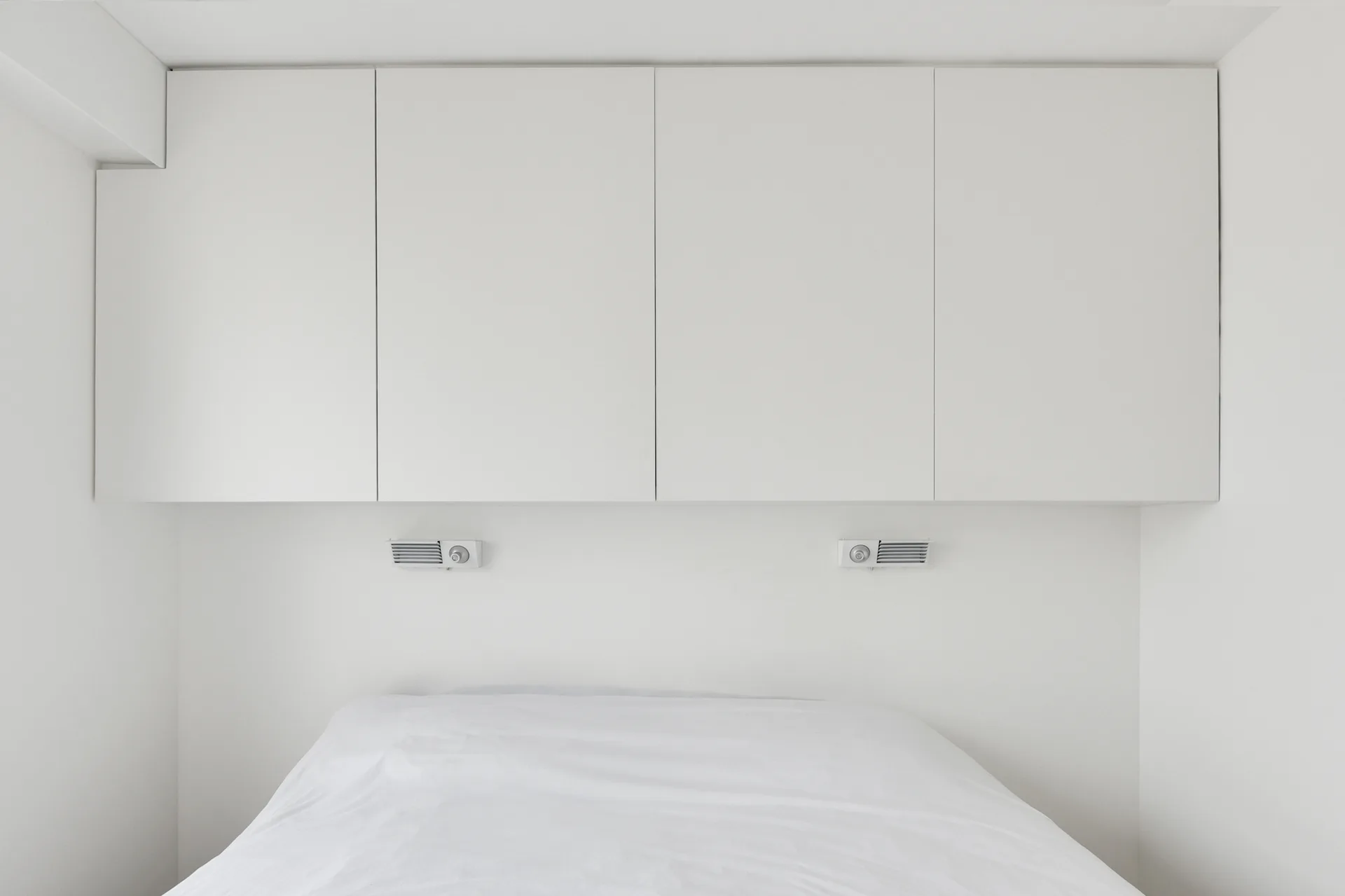 A minimalist white bedroom features a low bed positioned below seamless wall-mounted overhead cabinets and two small reading lights.