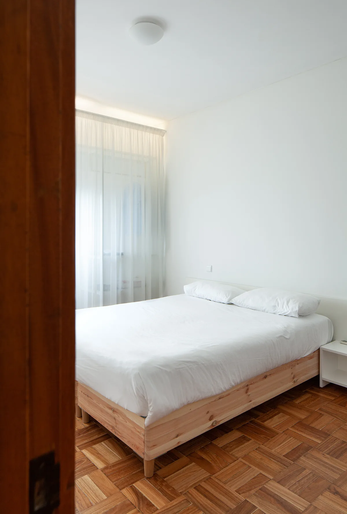 A minimalist bedroom featuring a natural wood bed frame with white bedding, sheer curtains, and parquet flooring viewed through a doorway.