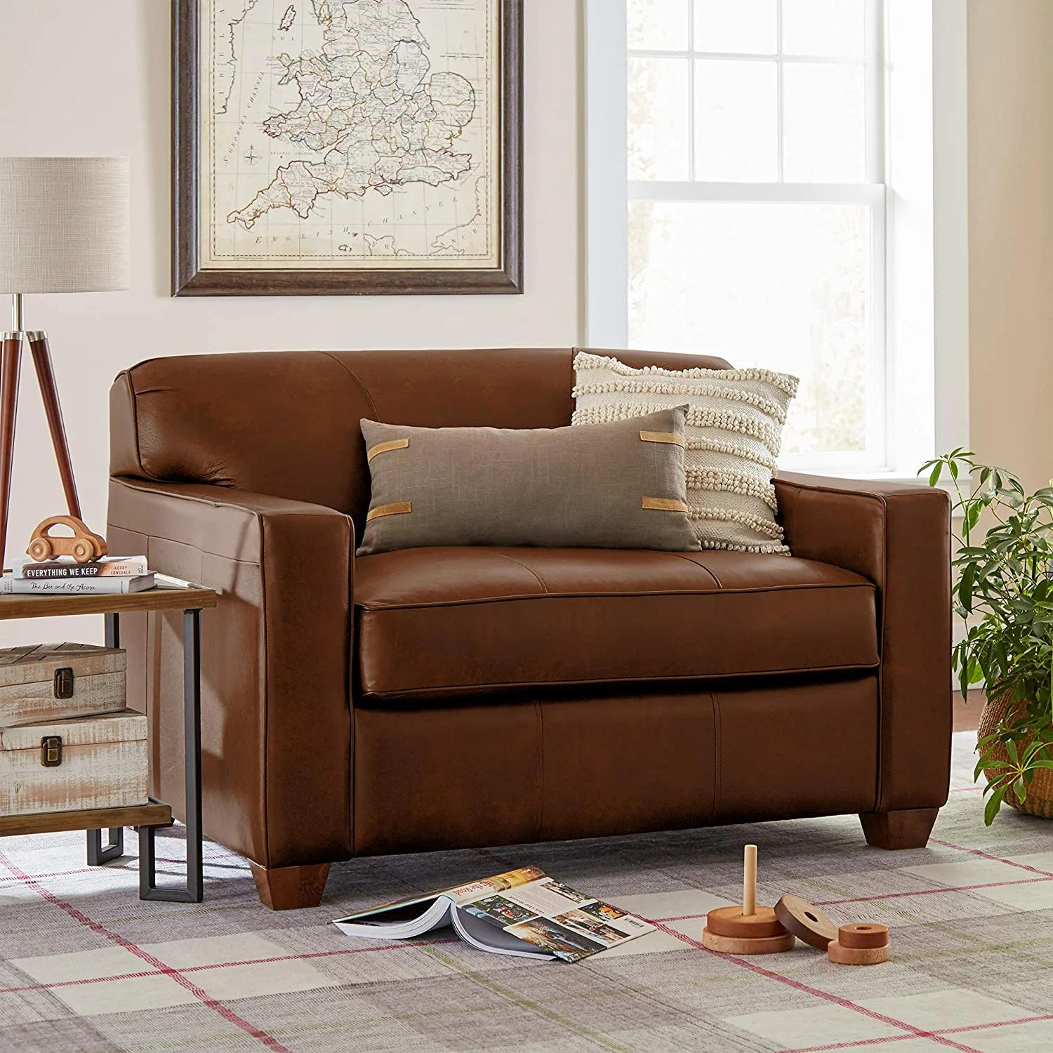 A brown leather armchair with decorative pillows sits on a plaid rug next to a small side table and a framed wall map.