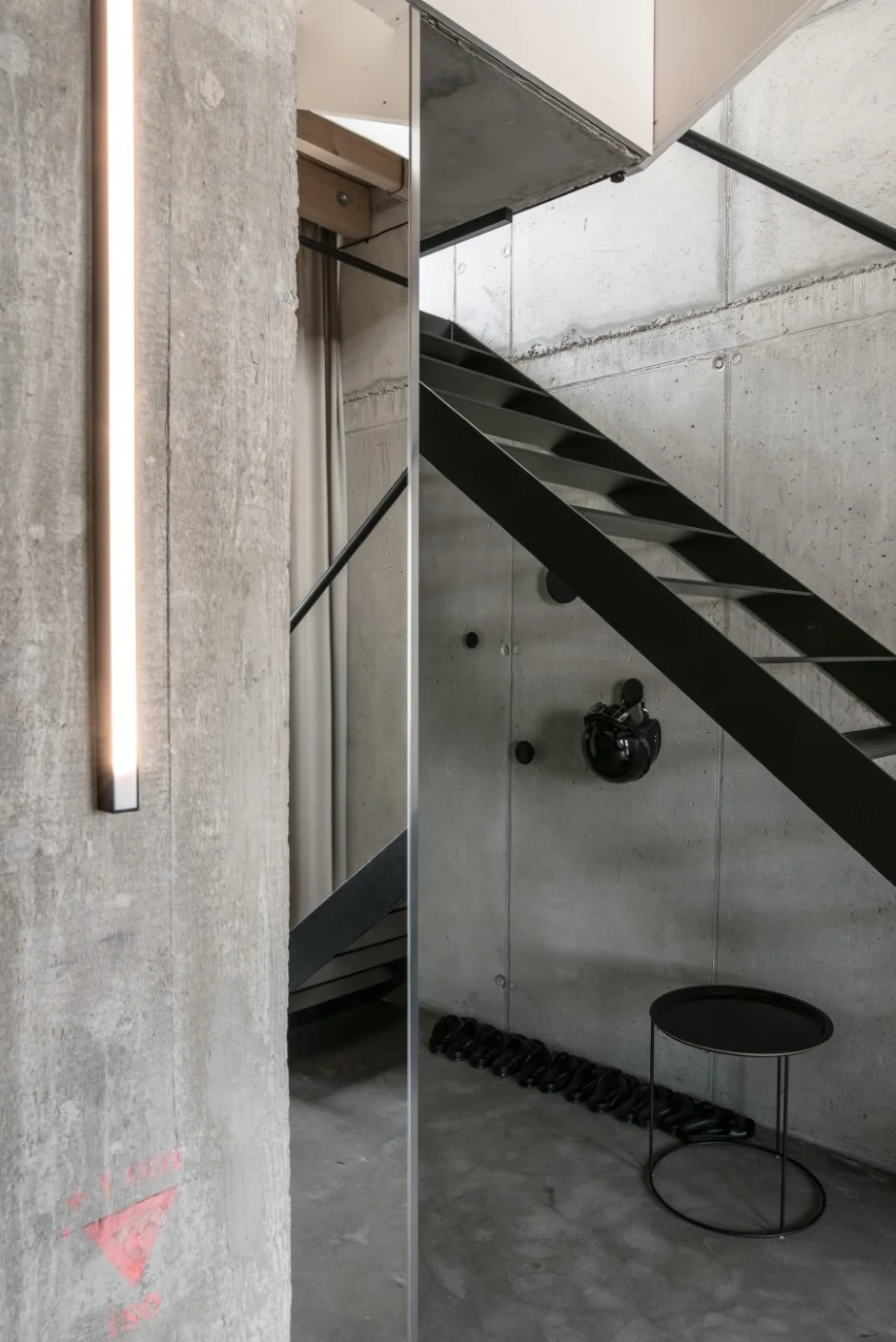 A black metal open-riser staircase stands against raw exposed concrete walls in an entryway featuring a vertical linear light and mirror.