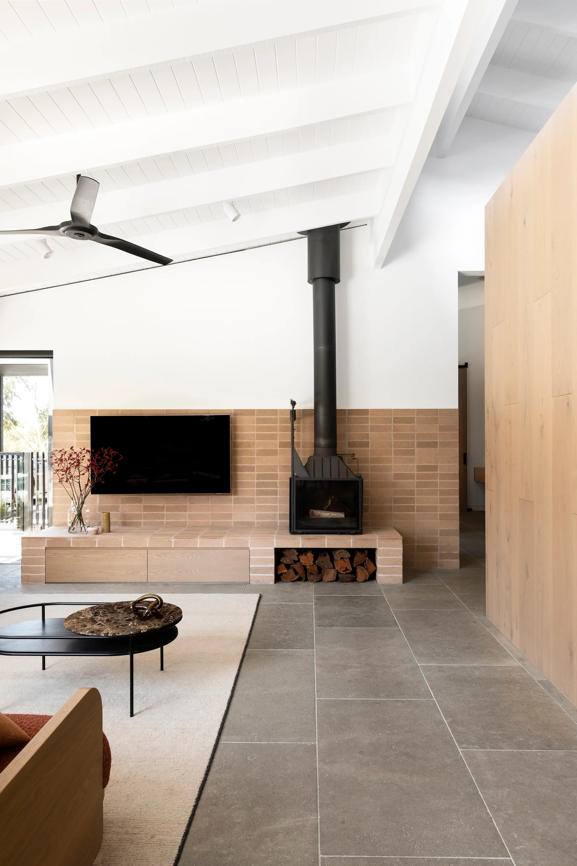 A modern living room features a black fireplace and wall-mounted television set against a low terracotta brick wall with white beamed ceilings above.