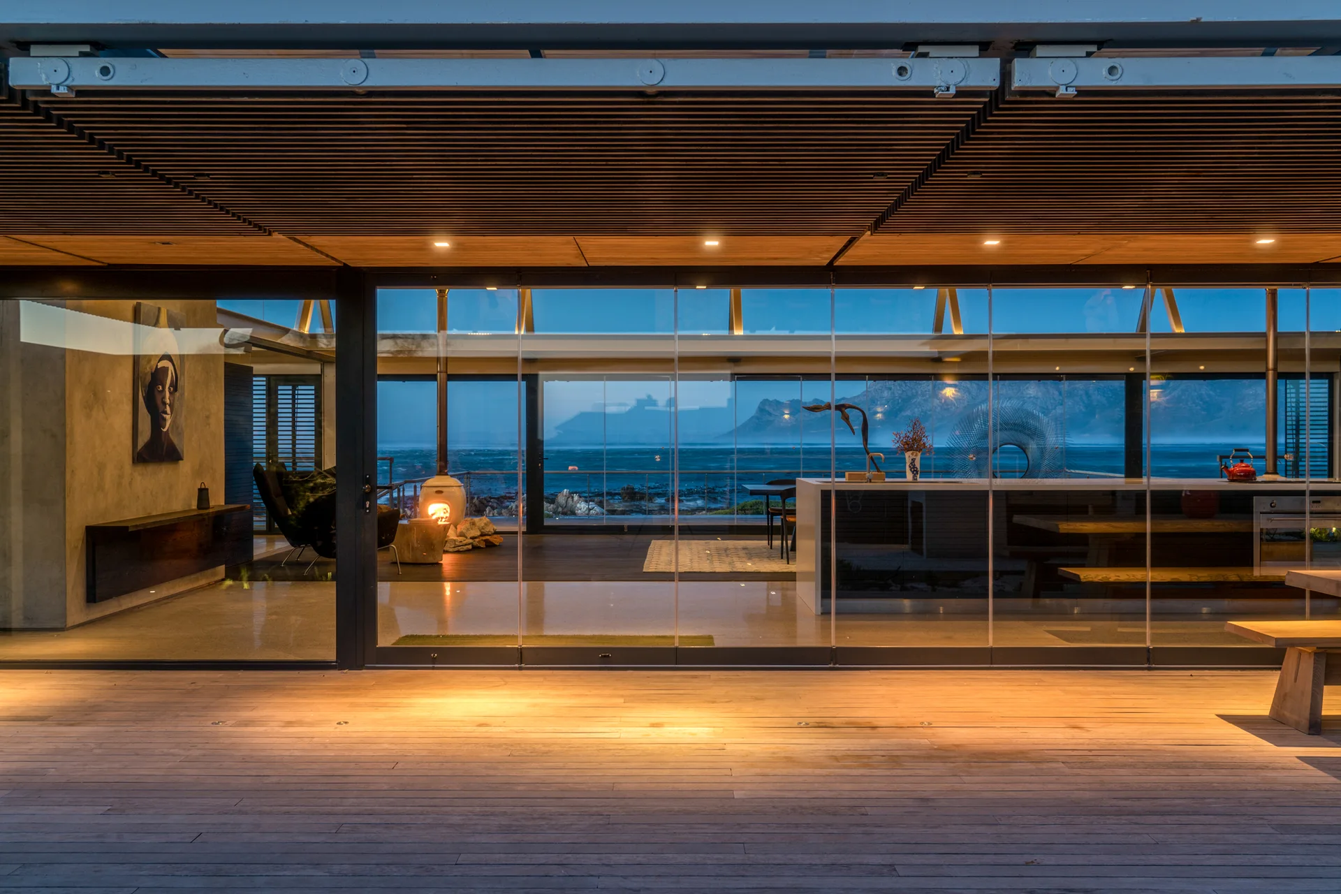 An open-plan coastal home at dusk featuring a wood-slatted ceiling, polished concrete floor, and floor-to-ceiling glass walls looking out to the sea.