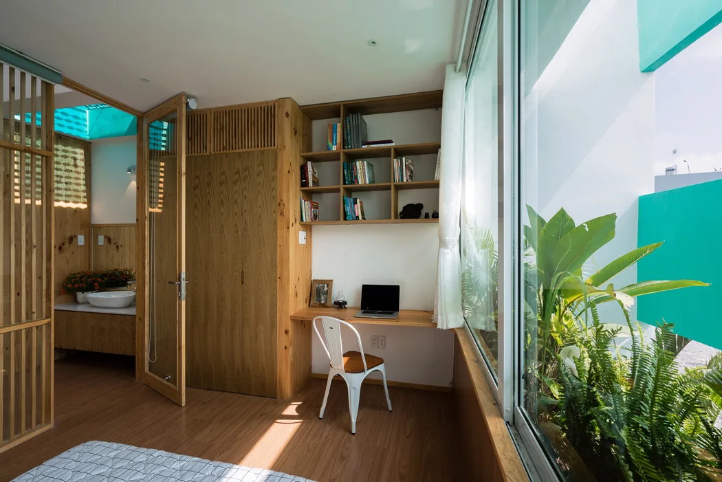 A modern bedroom features a built-in wooden desk and shelves next to a large glass window looking out at leafy green plants.