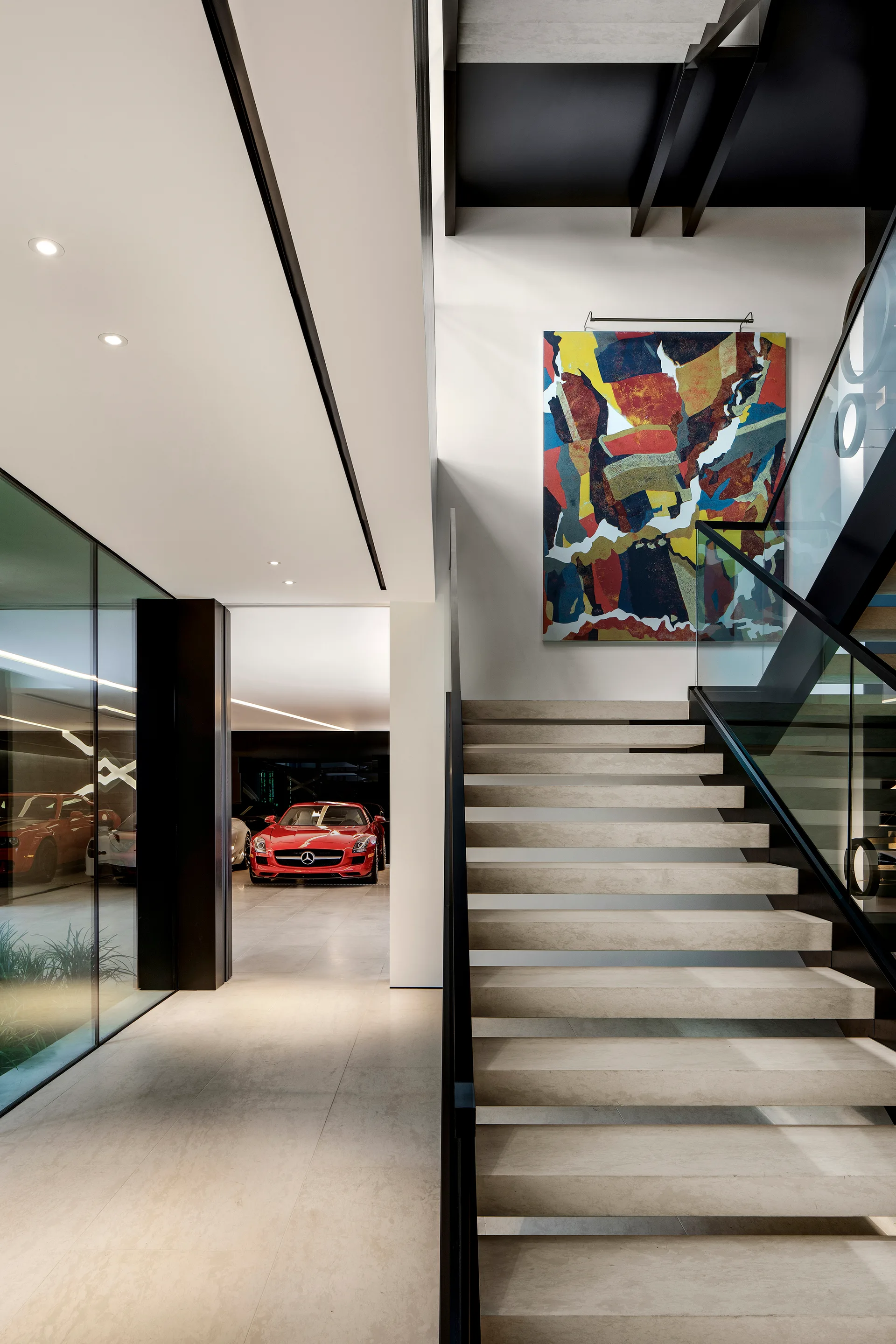 A modern stone staircase with a glass railing stands next to a clear glass partition showing a red sports car in a garage.