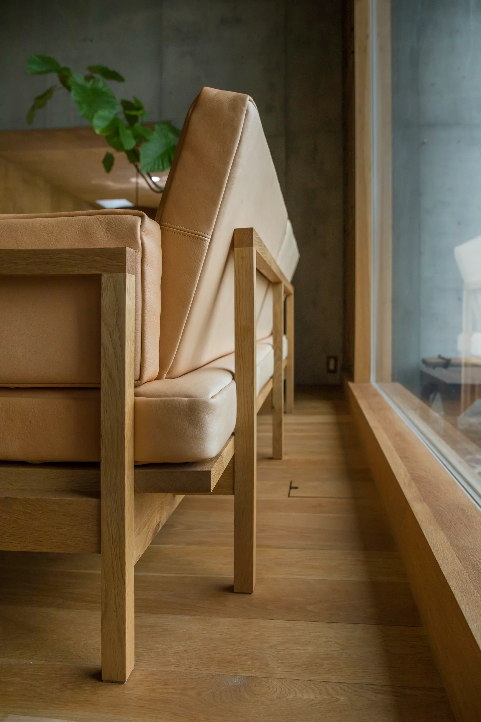 A close-up side view of a beige leather sofa with an exposed light oak wood frame on wood flooring next to a large window.