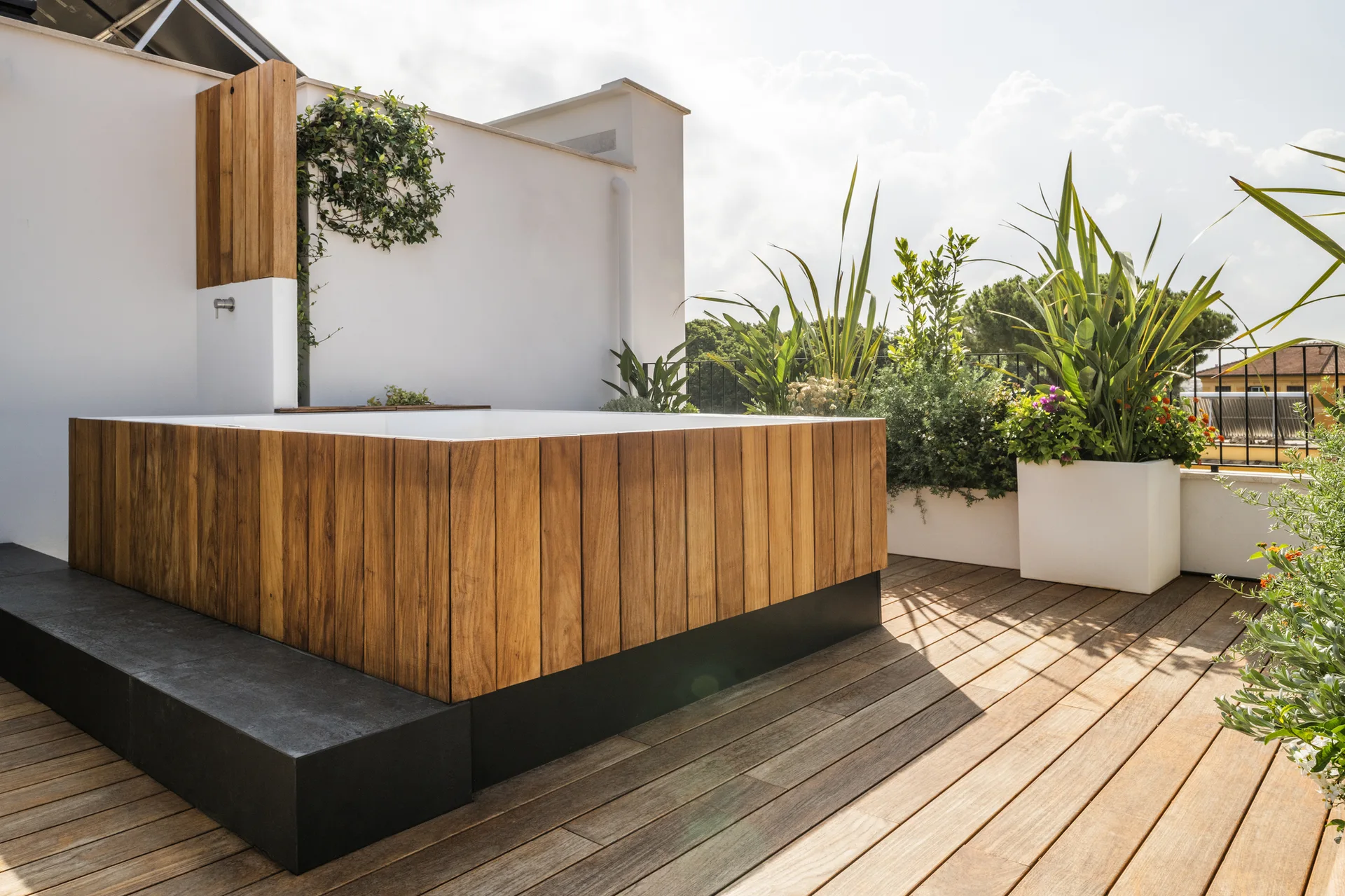 A rectangular hot tub clad in vertical wood slats sits on a rooftop terrace deck with white planters, green plants, and a black step.