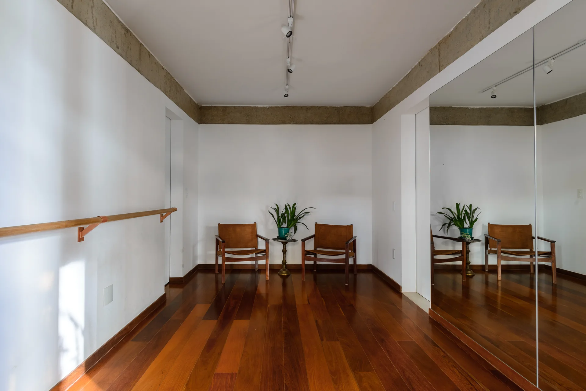 A minimalist interior space featuring two brown leather chairs, a small brass side table, polished hardwood floors, and a large wall-mounted mirror.