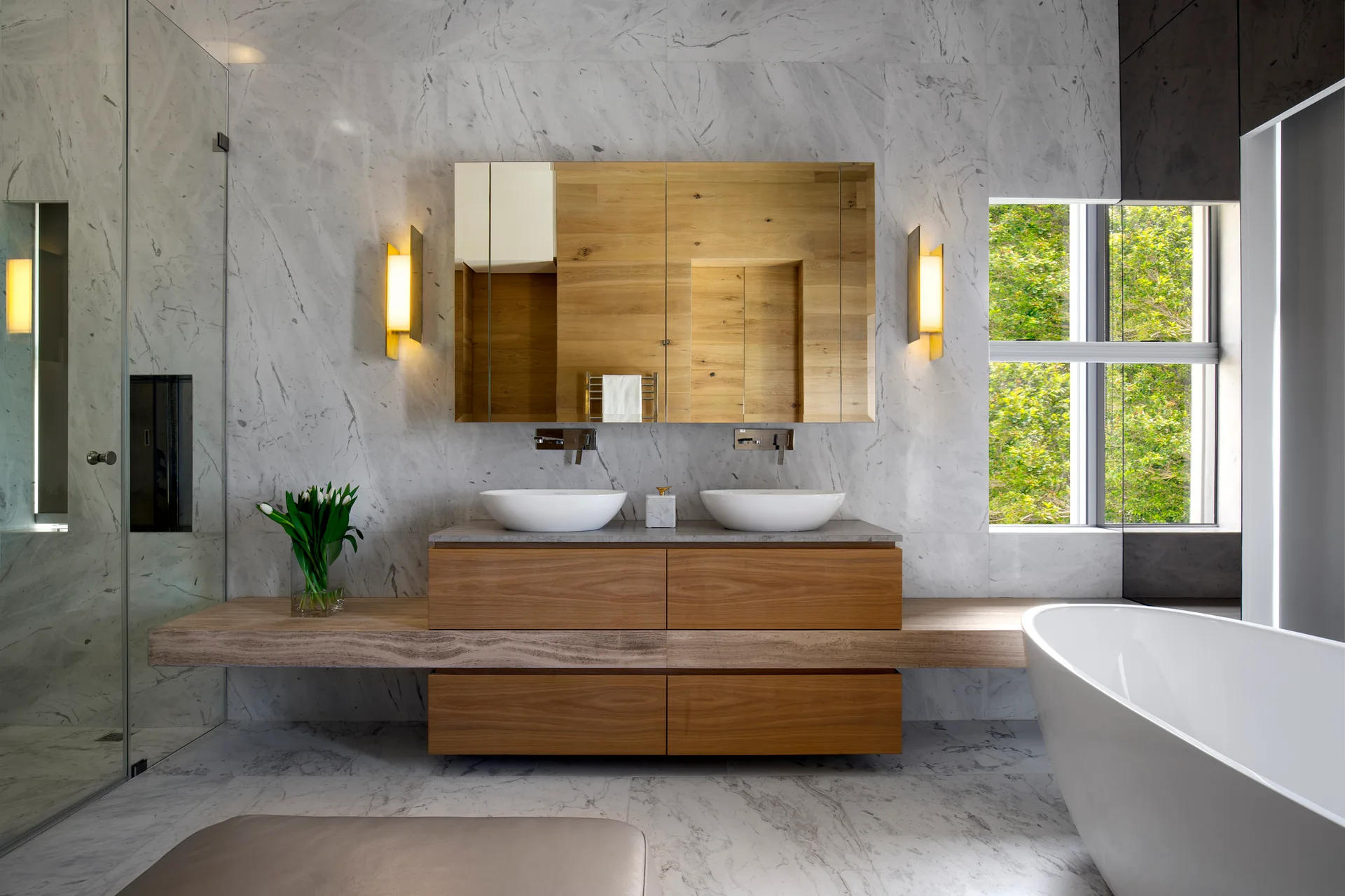 A modern bathroom with a wooden double vanity, two vessel sinks, a matching wooden wall cabinet, marble wall tiles, and a freestanding tub.