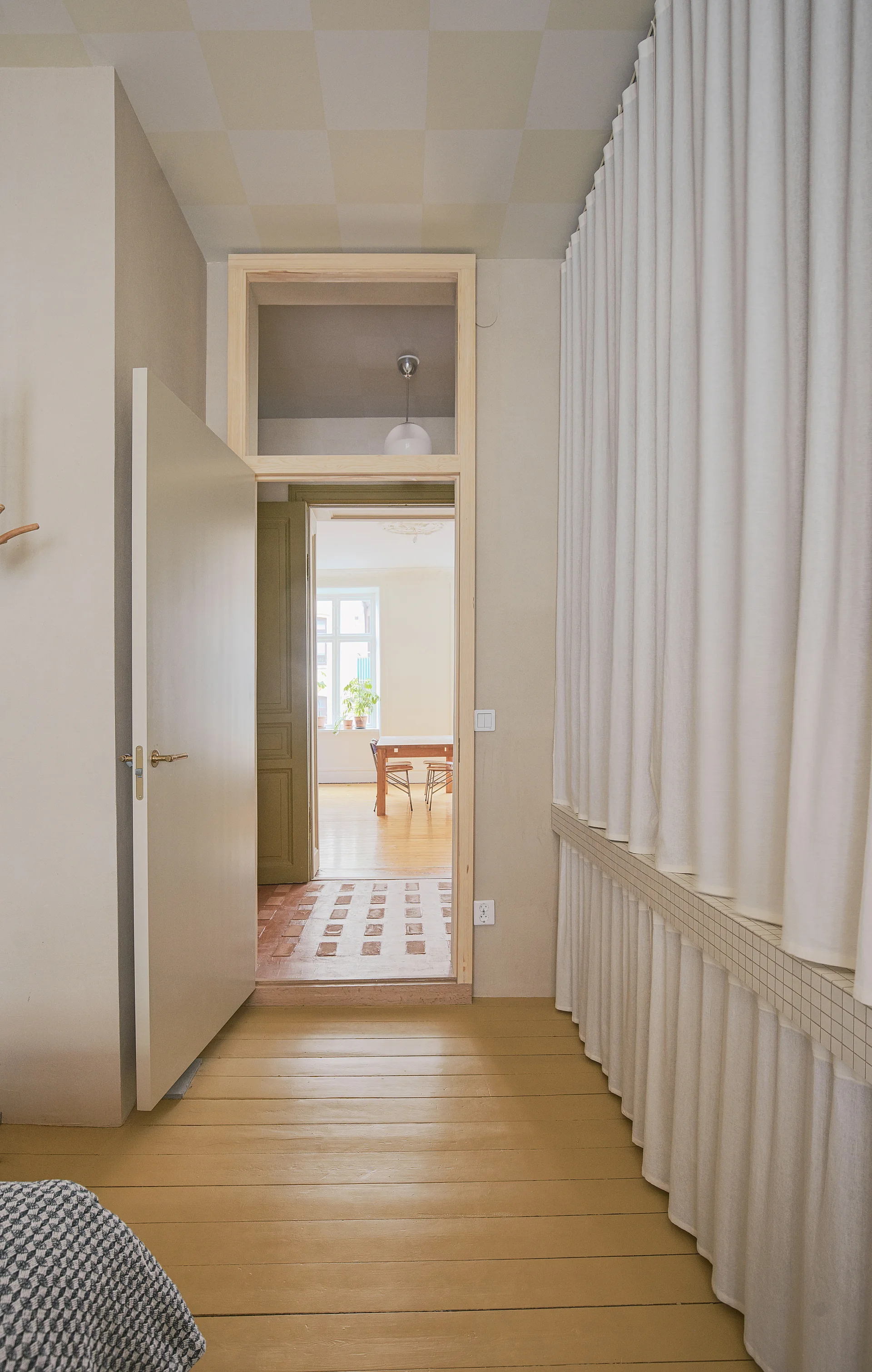 An open doorway with a transom window connects a hallway featuring painted yellow wood floors to a brightly lit dining area.