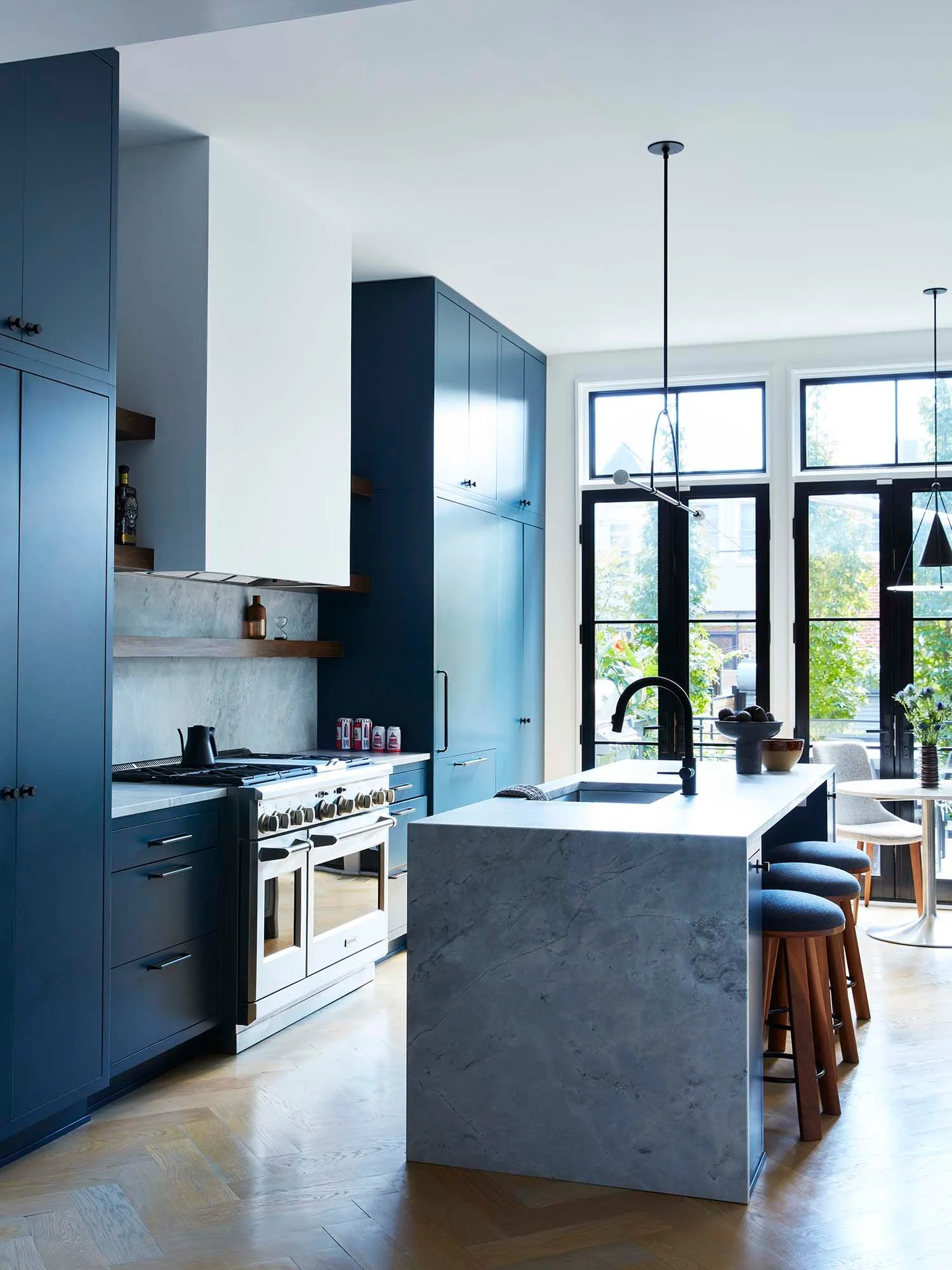 A modern kitchen featuring floor-to-ceiling navy blue cabinets, a grey stone waterfall island with wooden stools, and herringbone wood floors.