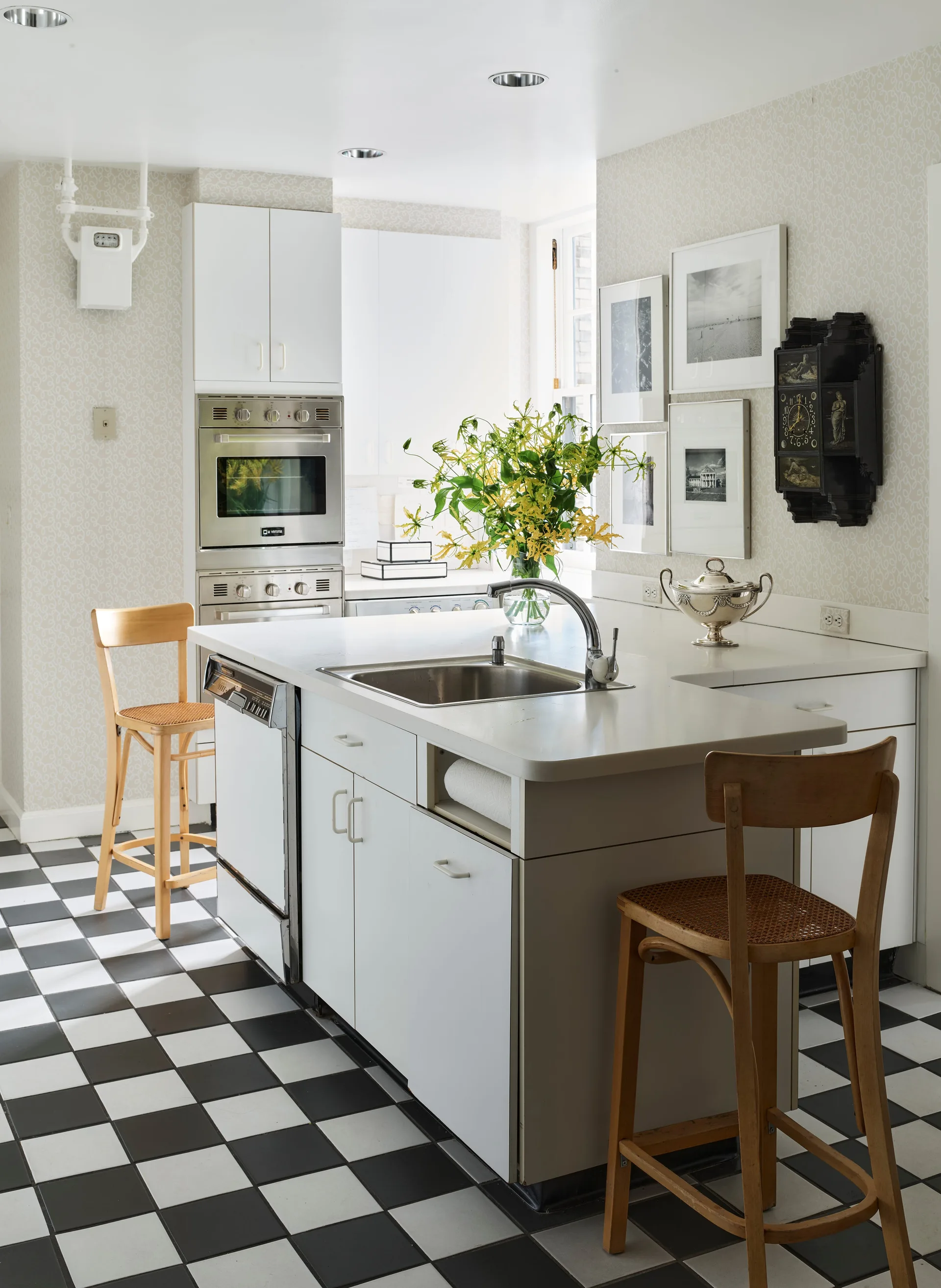 A kitchen featuring a white island with a sink, wooden bar stools, black-and-white checkered floor tiles, and delicate patterned wallpaper.