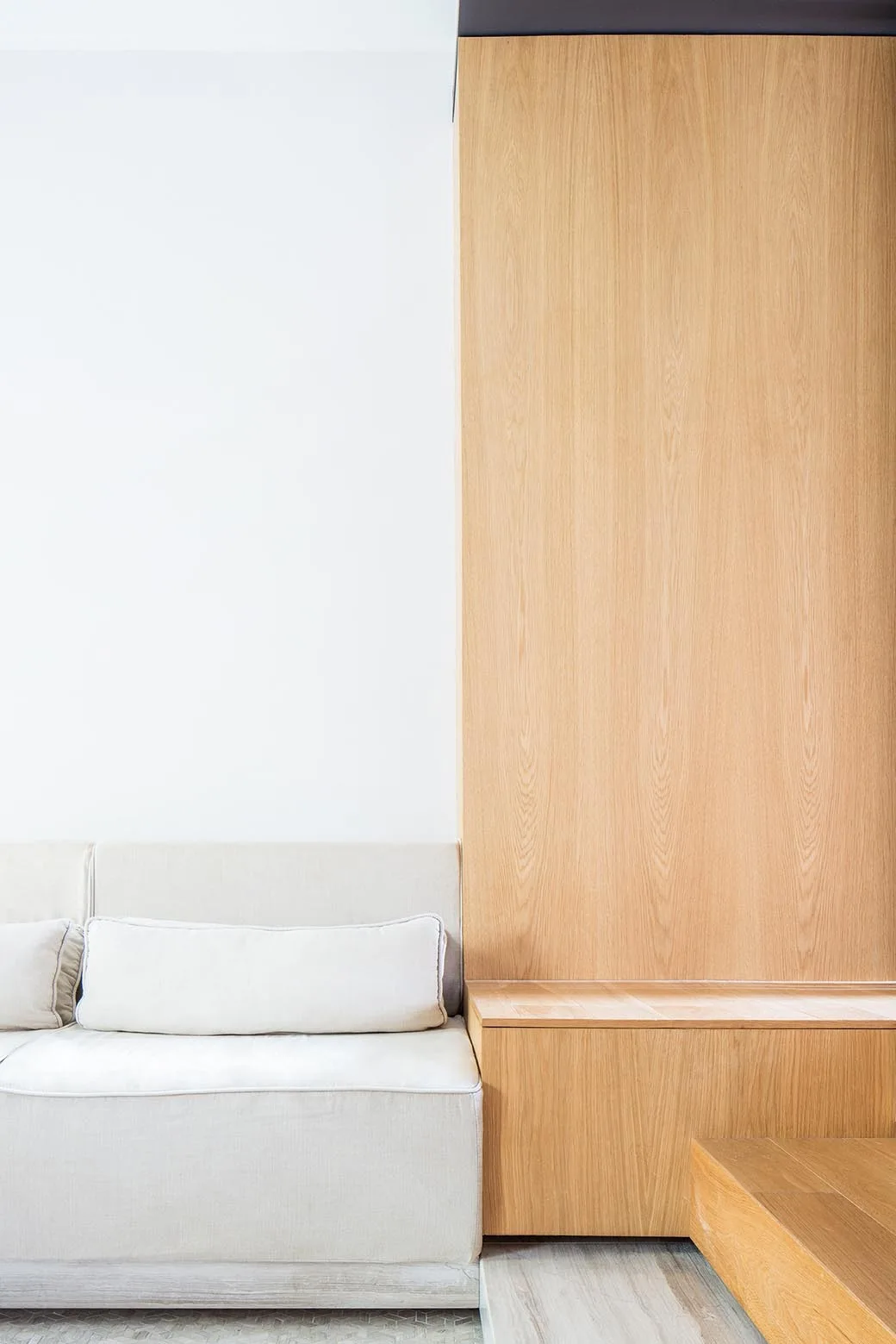 A cream fabric sofa with a rectangular throw pillow sits next to a large vertical light wood wall panel and an integrated wooden bench.