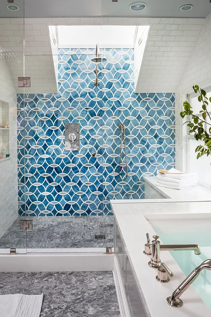 A bathroom features a glass shower enclosure with patterned blue tiles under a skylight next to a white bathtub with chrome fixtures.