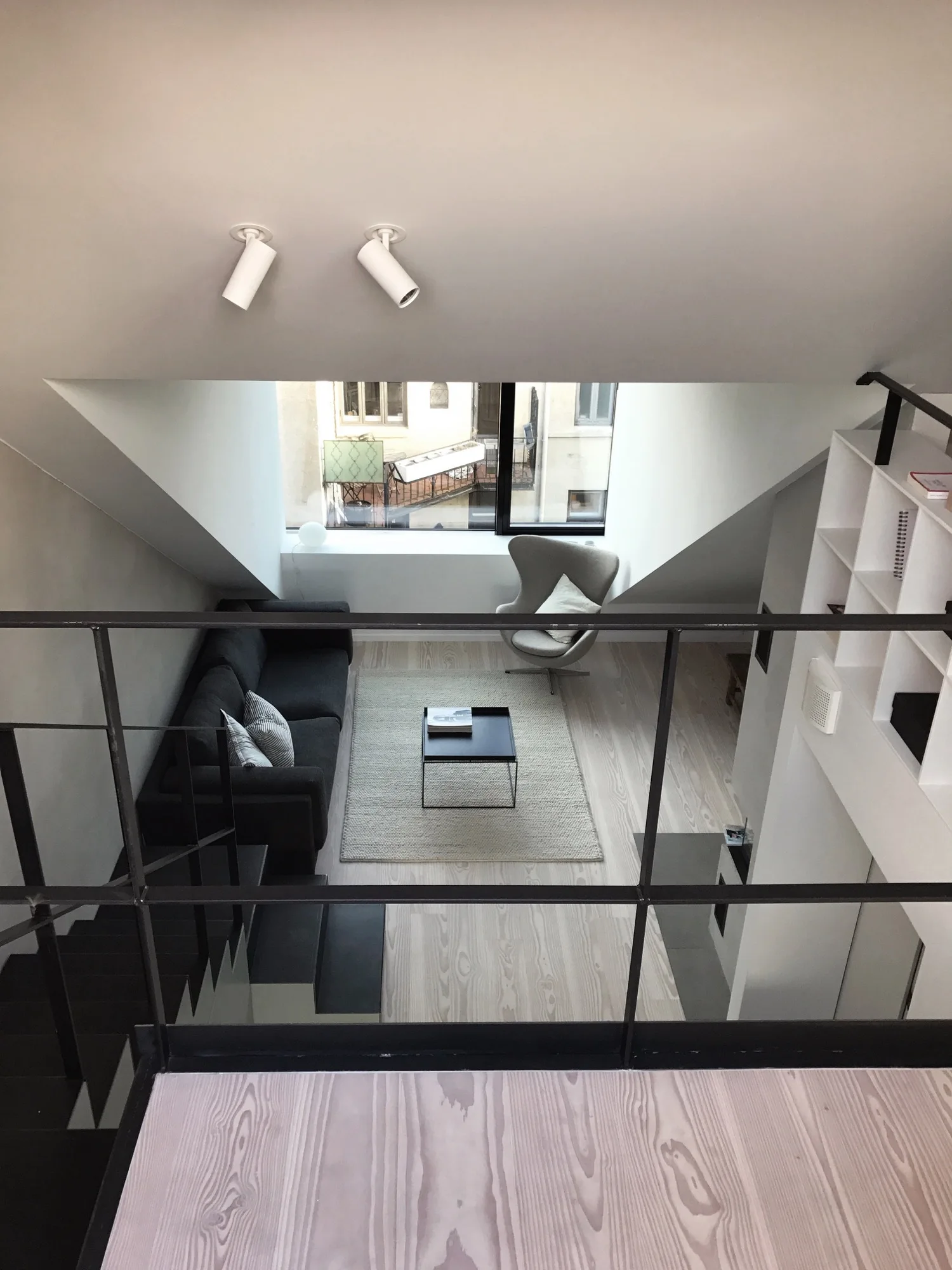 An elevated view looking down into a minimalist living room with a dark sofa, light wood floors, a grey lounge chair, and a large window.