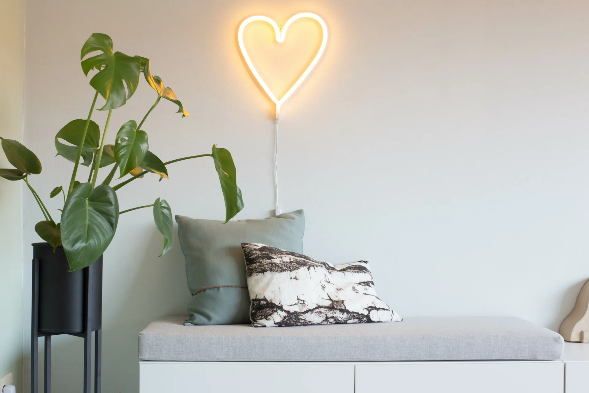 A grey upholstered bench with decorative pillows sits next to a potted monstera plant beneath a glowing heart-shaped neon wall light.