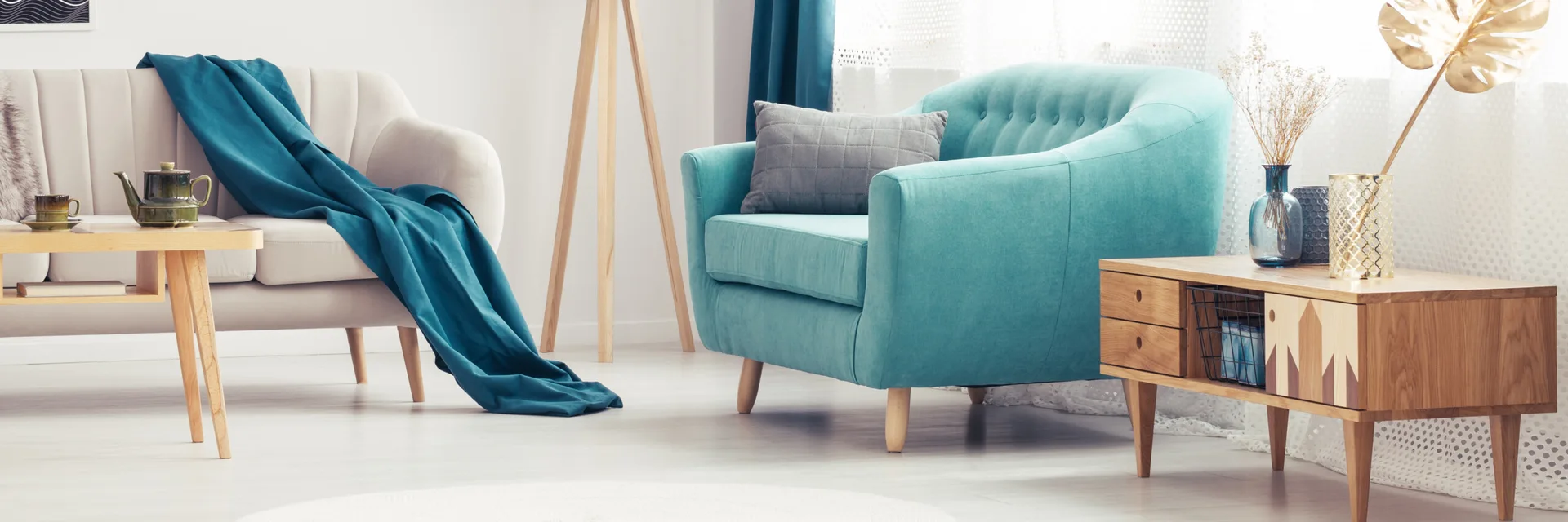 A teal tufted armchair stands next to a wooden mid-century modern side table and a white accent chair in a bright living room.