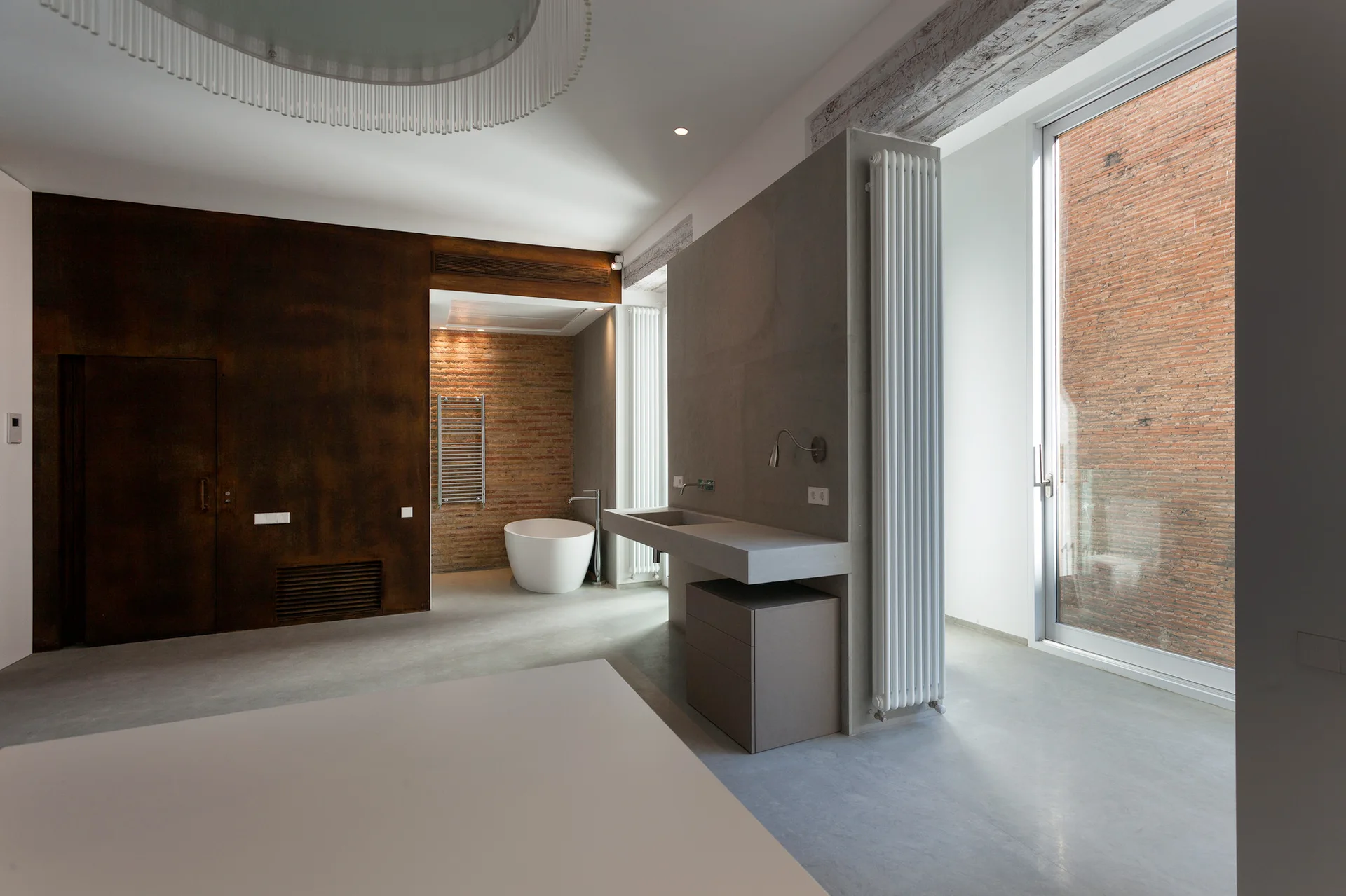 A modern loft bathroom featuring a long concrete vanity, a freestanding white bathtub against an exposed brick wall, and dark rusted metal wall paneling.