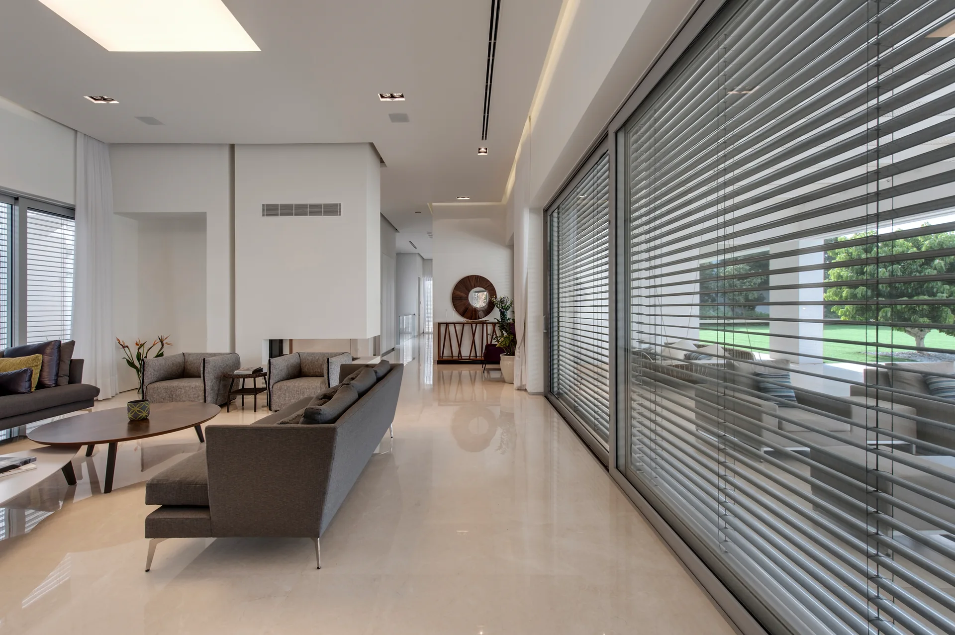 A spacious living room features gray sofas, an oval wood coffee table, and large windows with horizontal venetian blinds over polished stone floors.