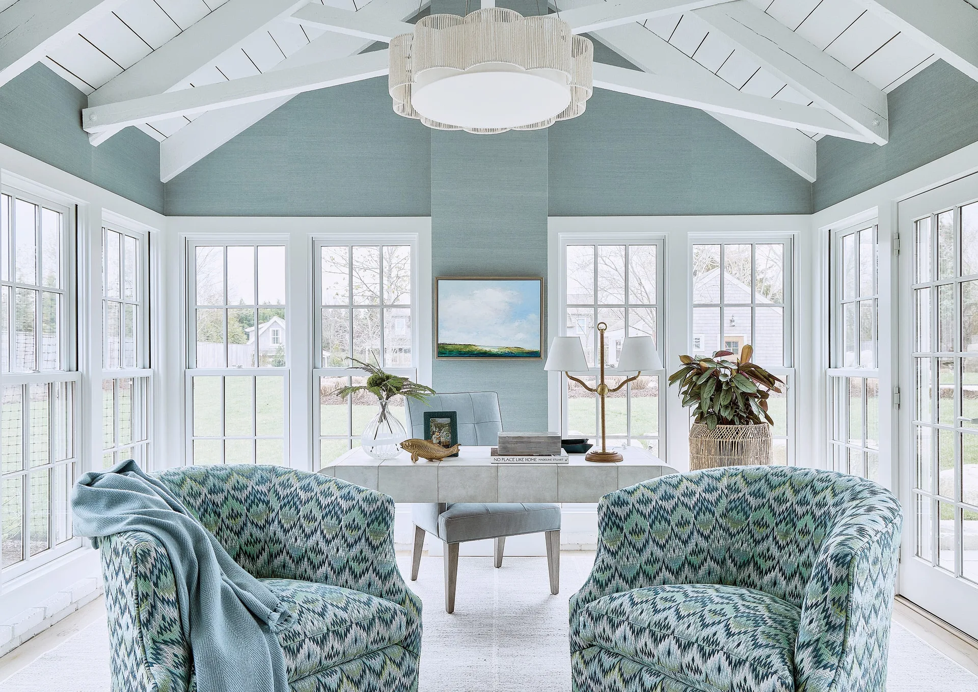 A bright home office features two blue patterned armchairs, a white writing desk, and blue textured wallpaper under a vaulted beamed ceiling.