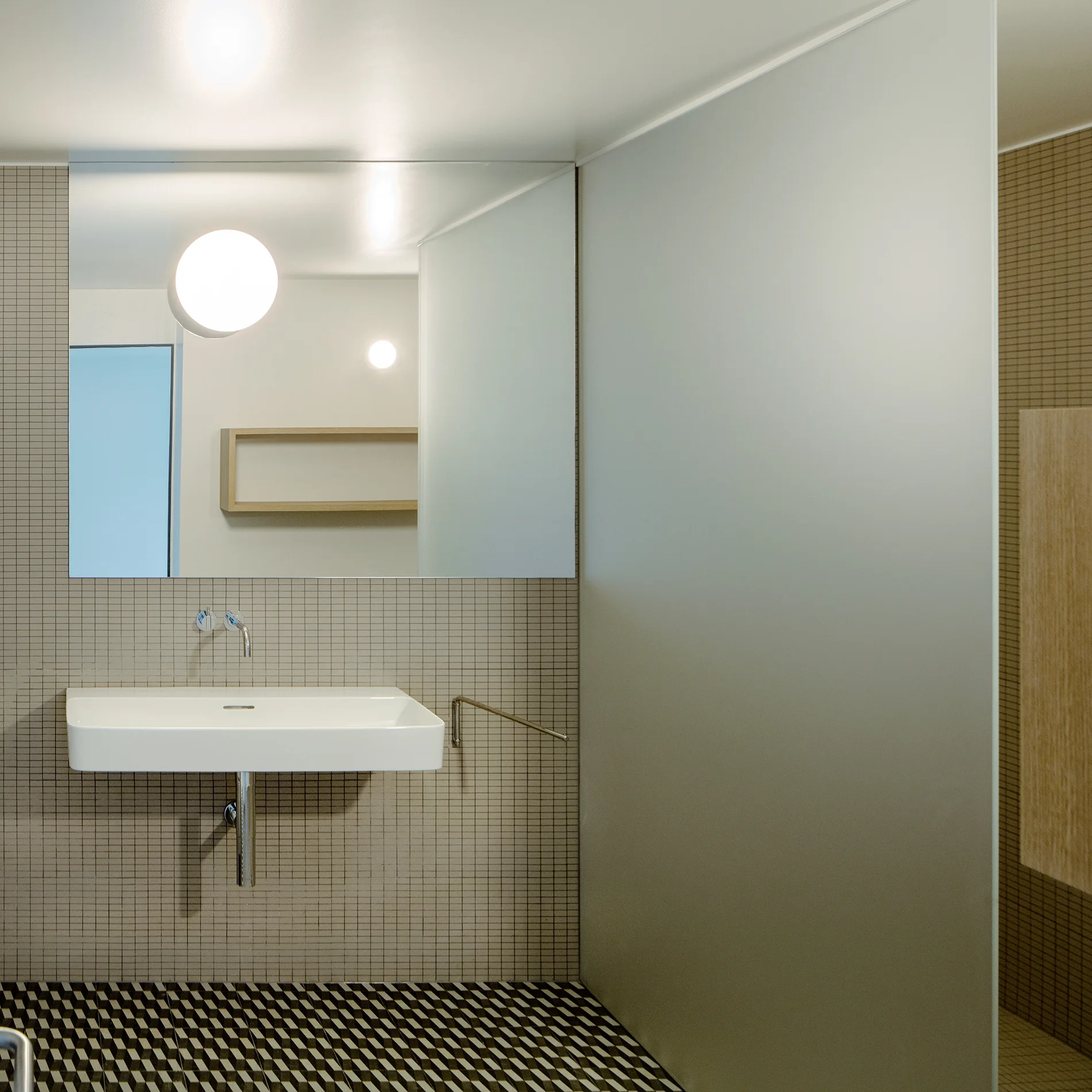 A minimalist bathroom featuring a wall-mounted ceramic sink, beige mosaic wall tiles, a frosted glass partition, and a geometric tiled floor.